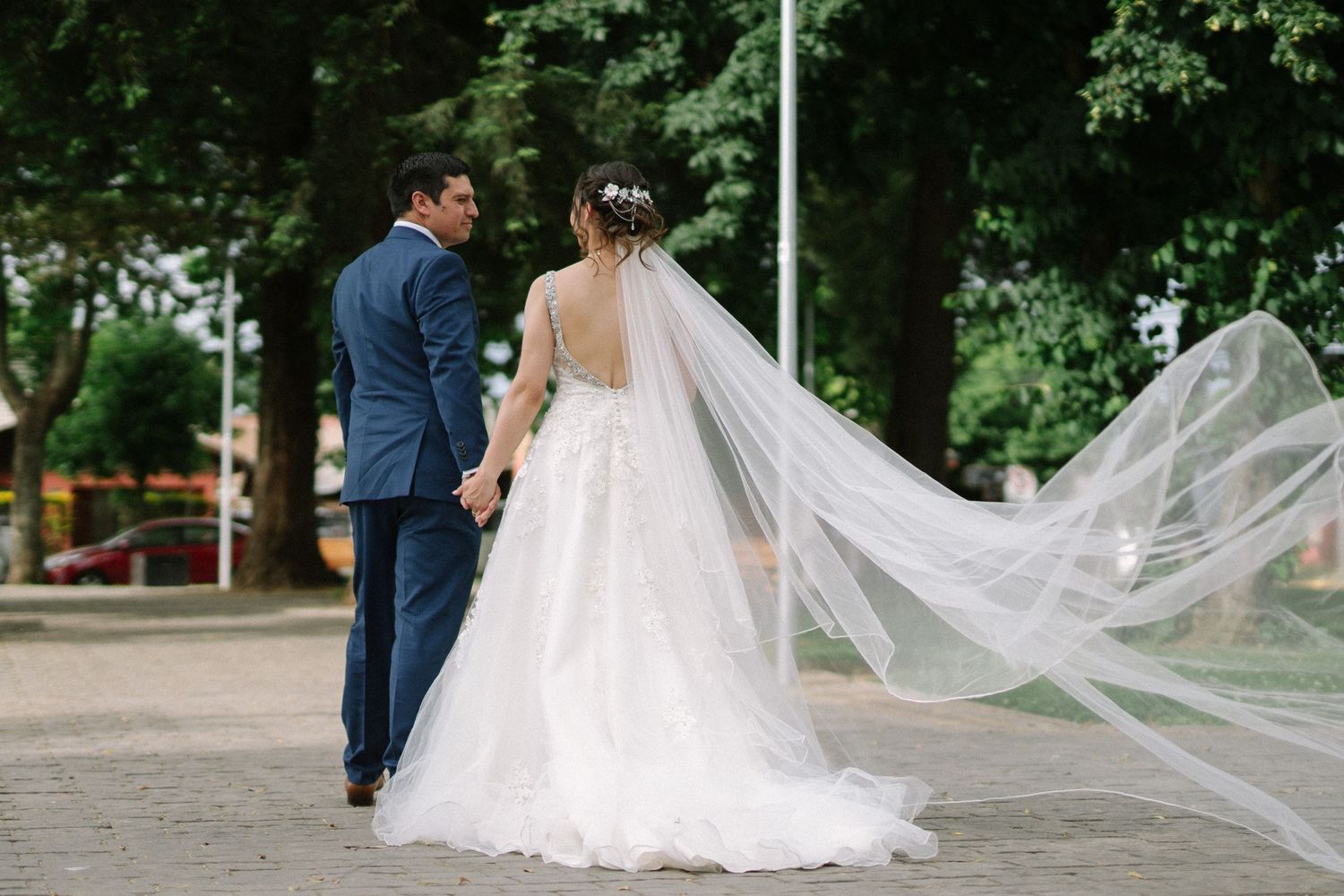 Bride's flowing veil captures movement as couple walks through park.
