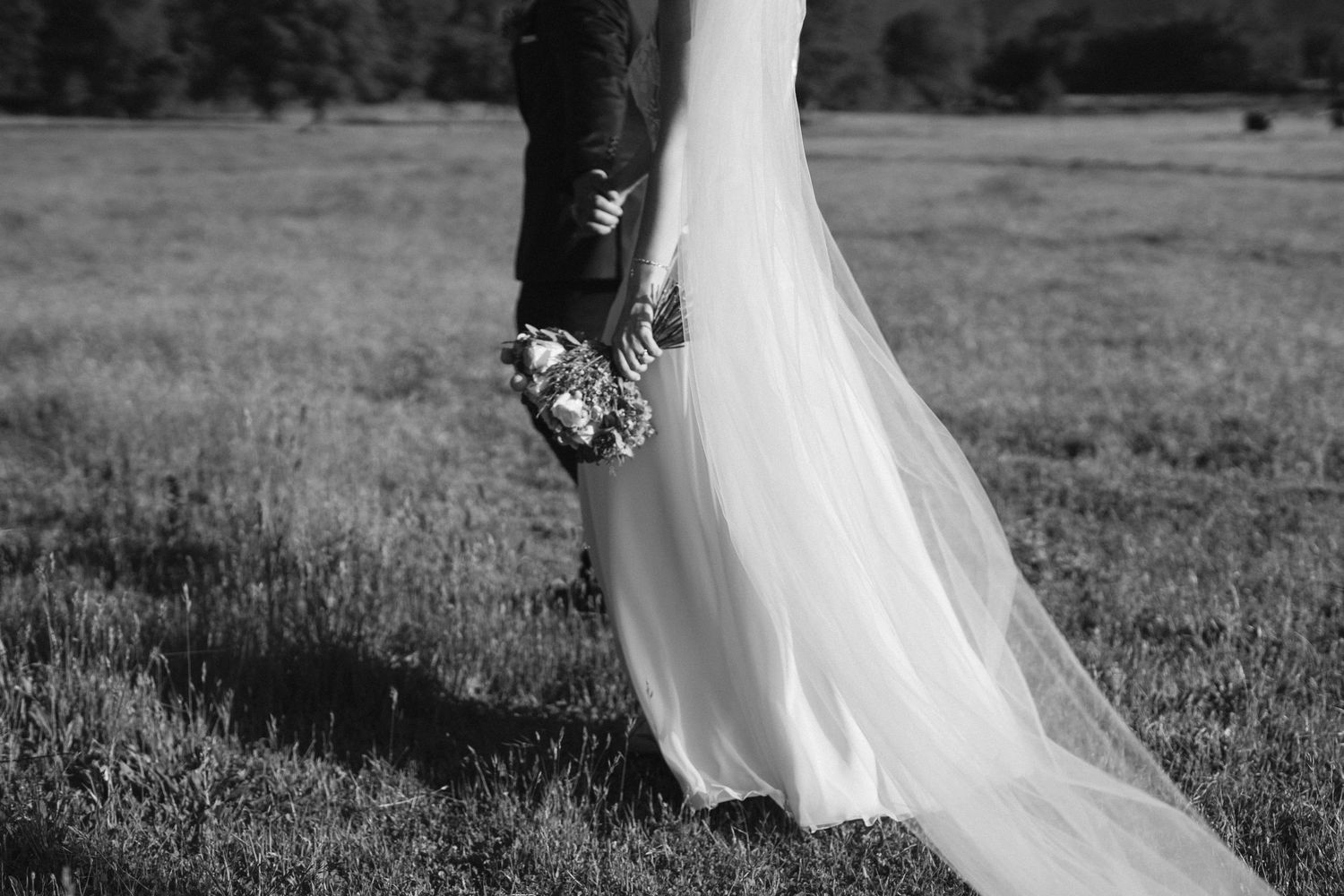 Black and white image of a bride's flowing dress and bouquet against a pastoral field background.
