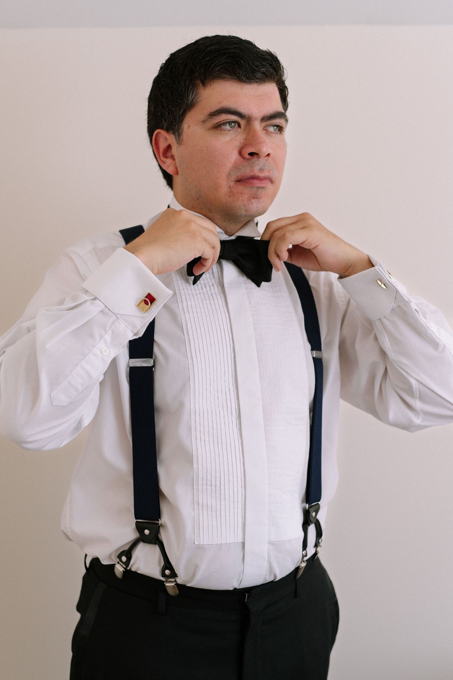Person adjusting black bow tie with white tuxedo shirt and suspenders.
