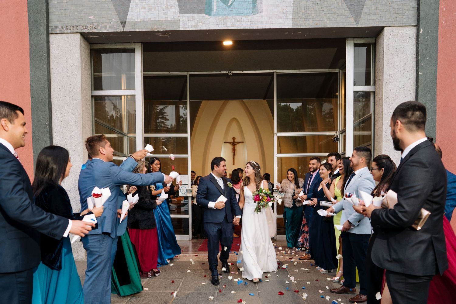Wedding guests throw confetti as couple exits church under modern architecture.