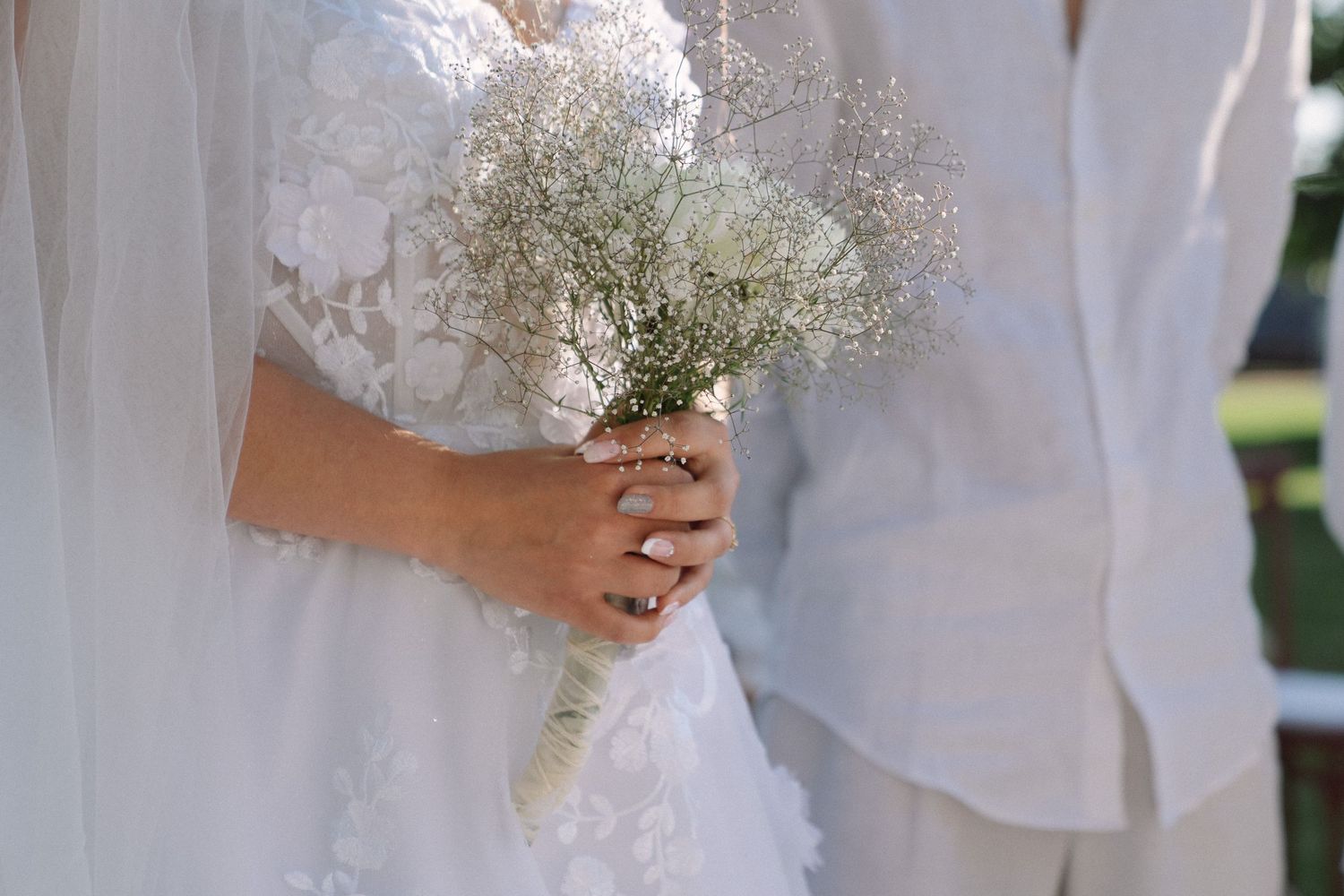 Bride in lace wedding dress holding a delicate white baby's breath bouquet during outdoor wedding ceremony.