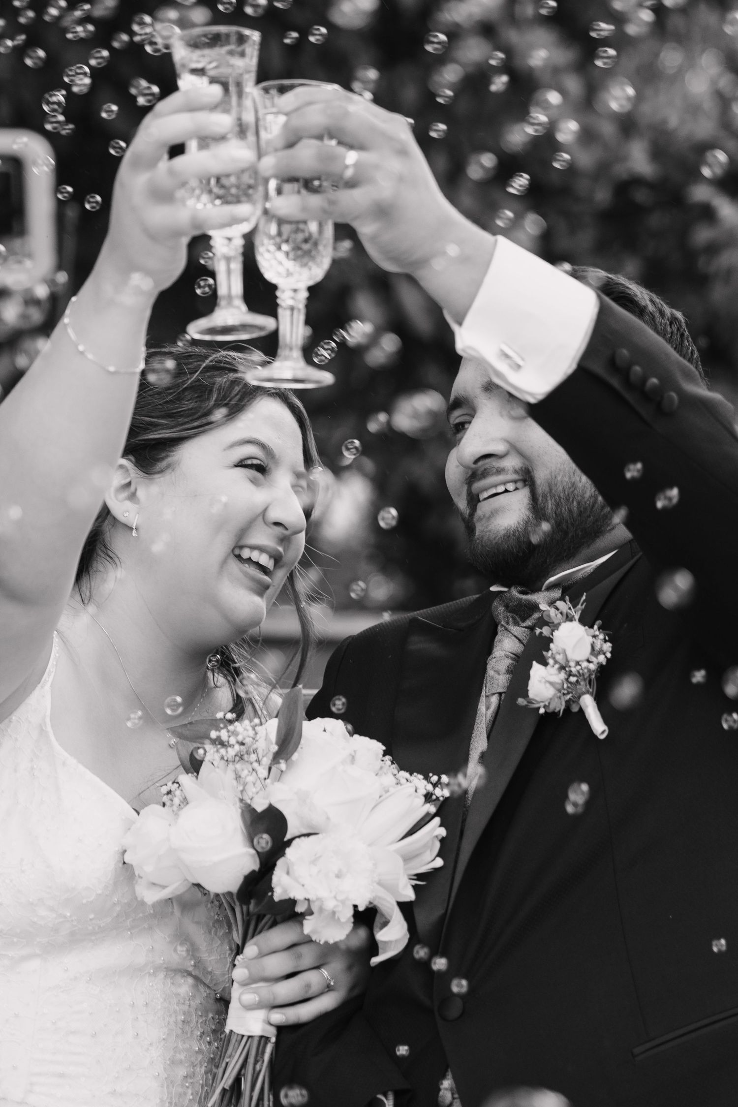 Black and white photo of happy newlyweds toasting champagne glasses surrounded by bubbles outdoors.