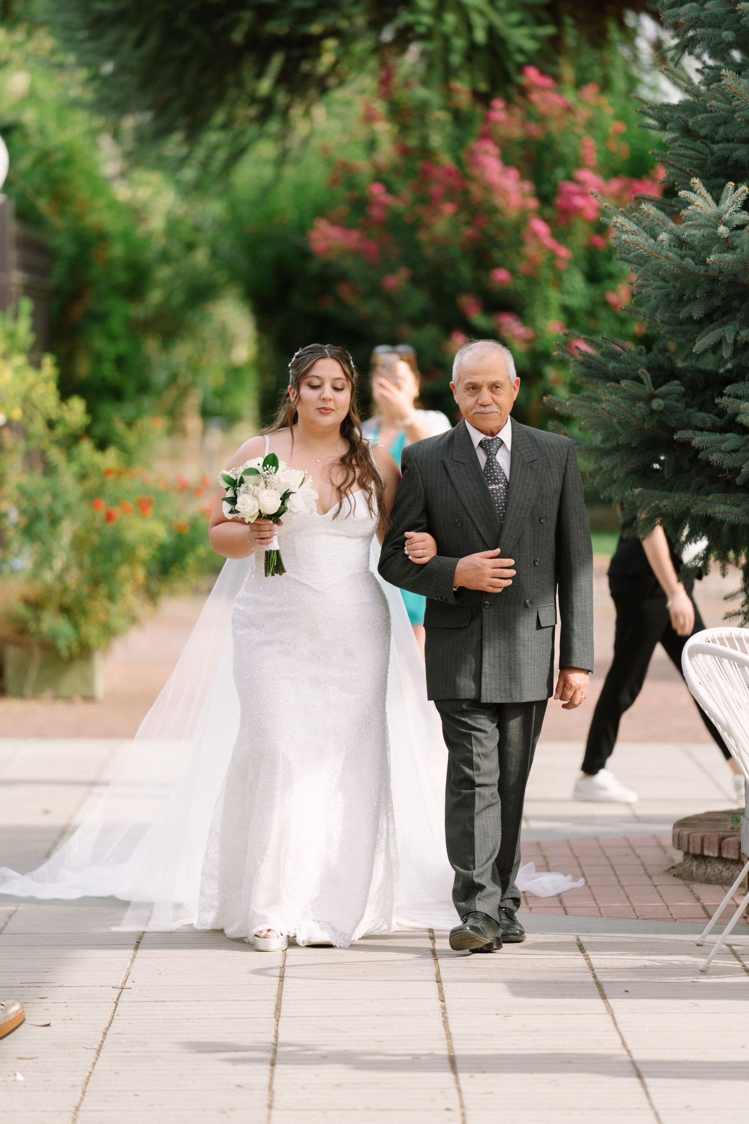 Bride in white mermaid gown holding bouquet walks down outdoor aisle with father among lush greenery.