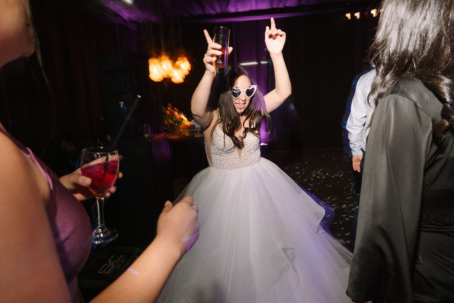 A bride in a white ballgown dances energetically under purple lighting at a reception.
