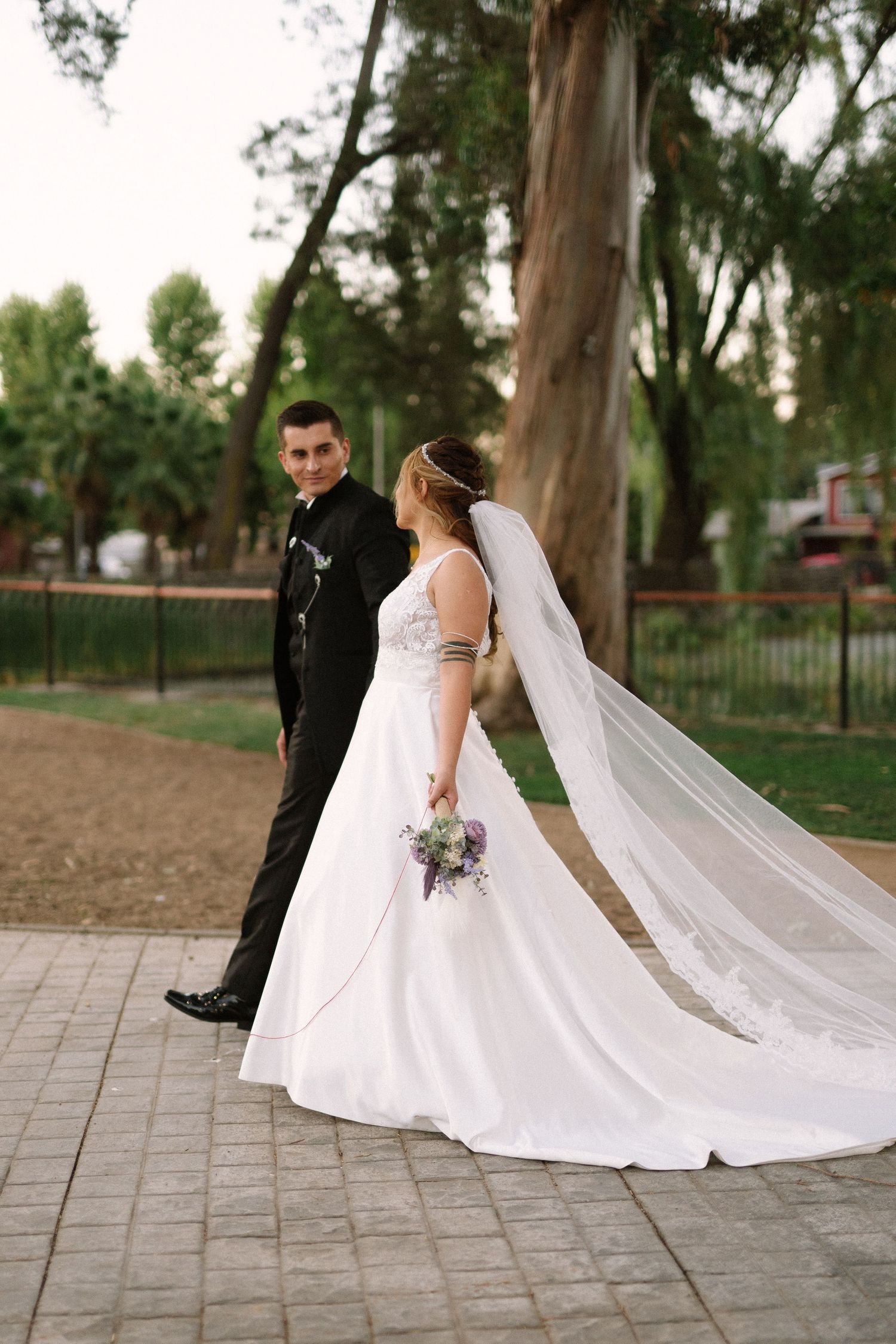 A couple in wedding attire walks along a paved path surrounded by tall trees in a park setting.
