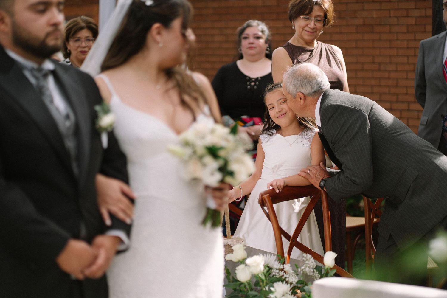 Groom kneeling to kiss a flower girl during an outdoor wedding ceremony.