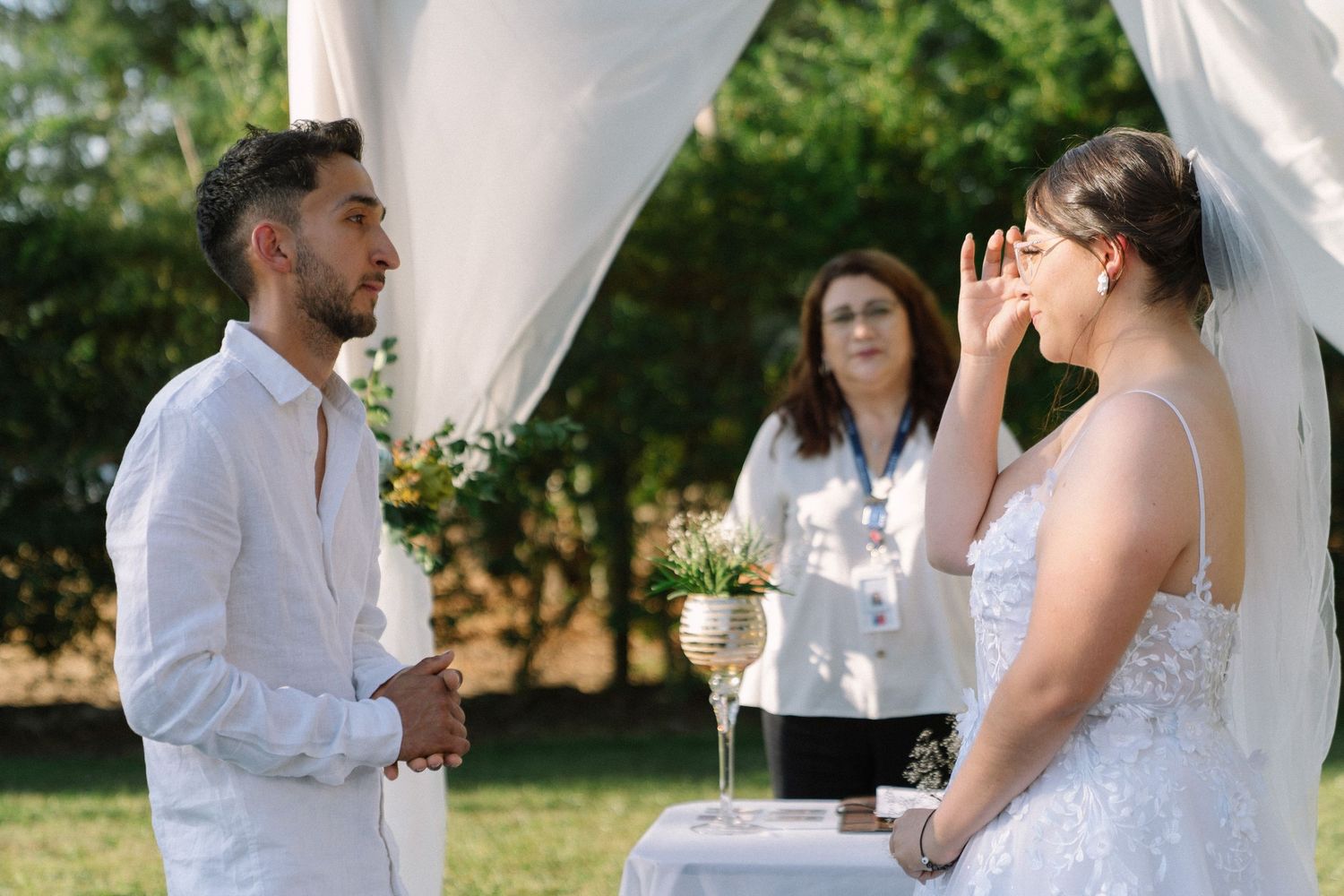 Emotional bride wiping tears as groom and officiant stand beneath white draped wedding arch outdoors.