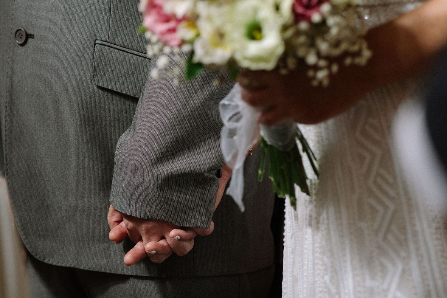 Hands clasped behind backs during a wedding ceremony beside a floral bouquet.