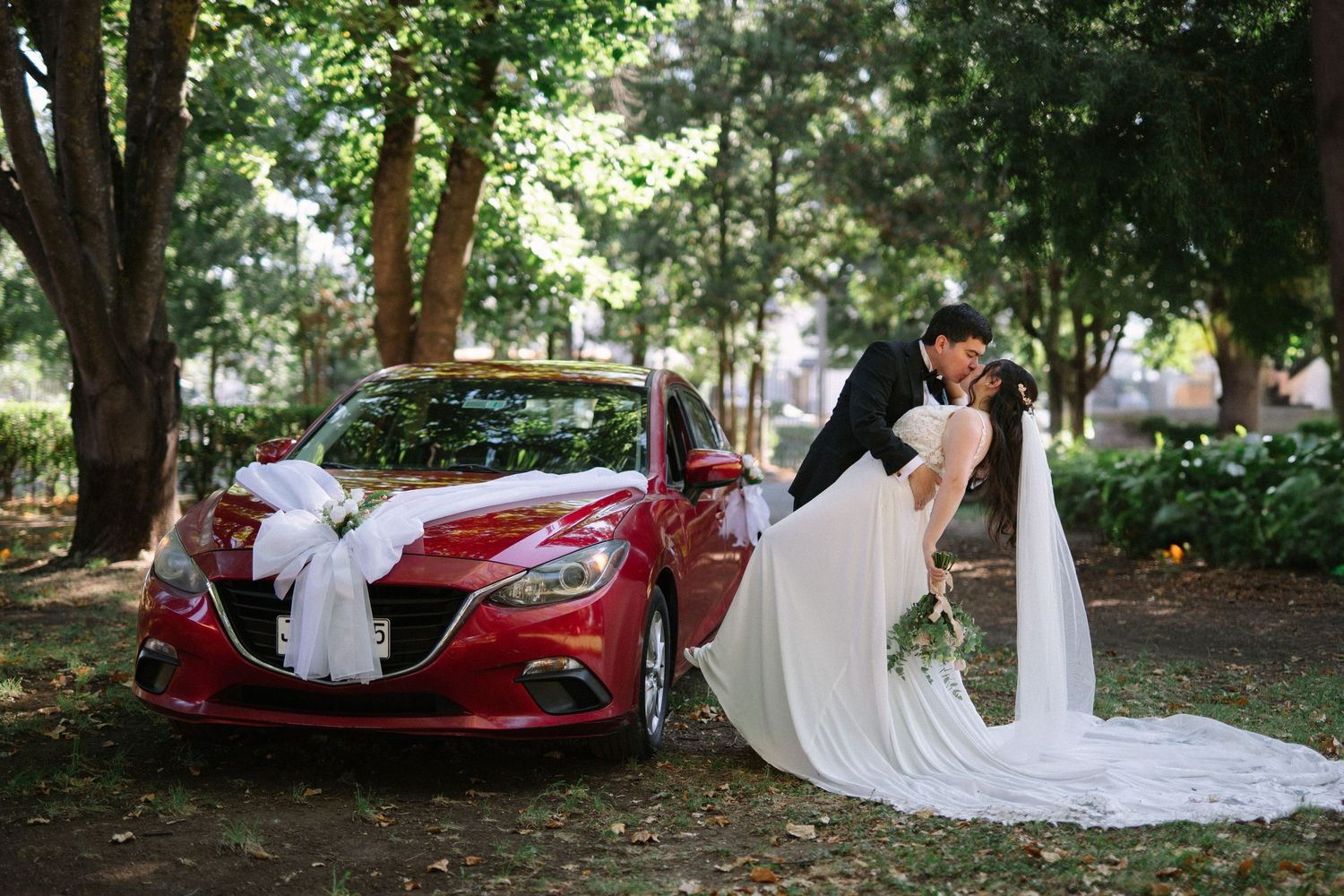 Wedding couple with flowing white dress veil standing by decorated red car.