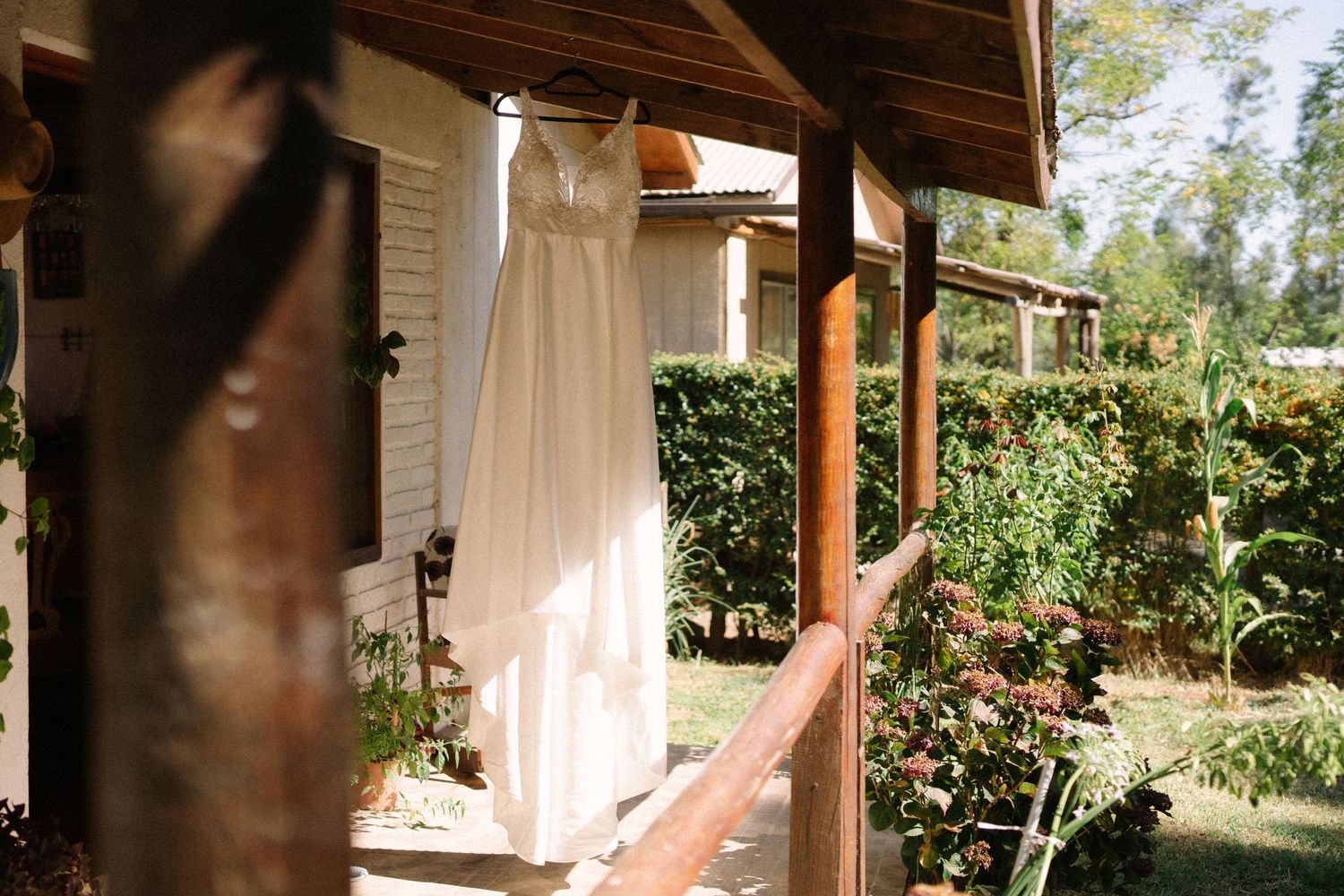 A white wedding dress hangs from a wooden porch beam with greenery in the background.
