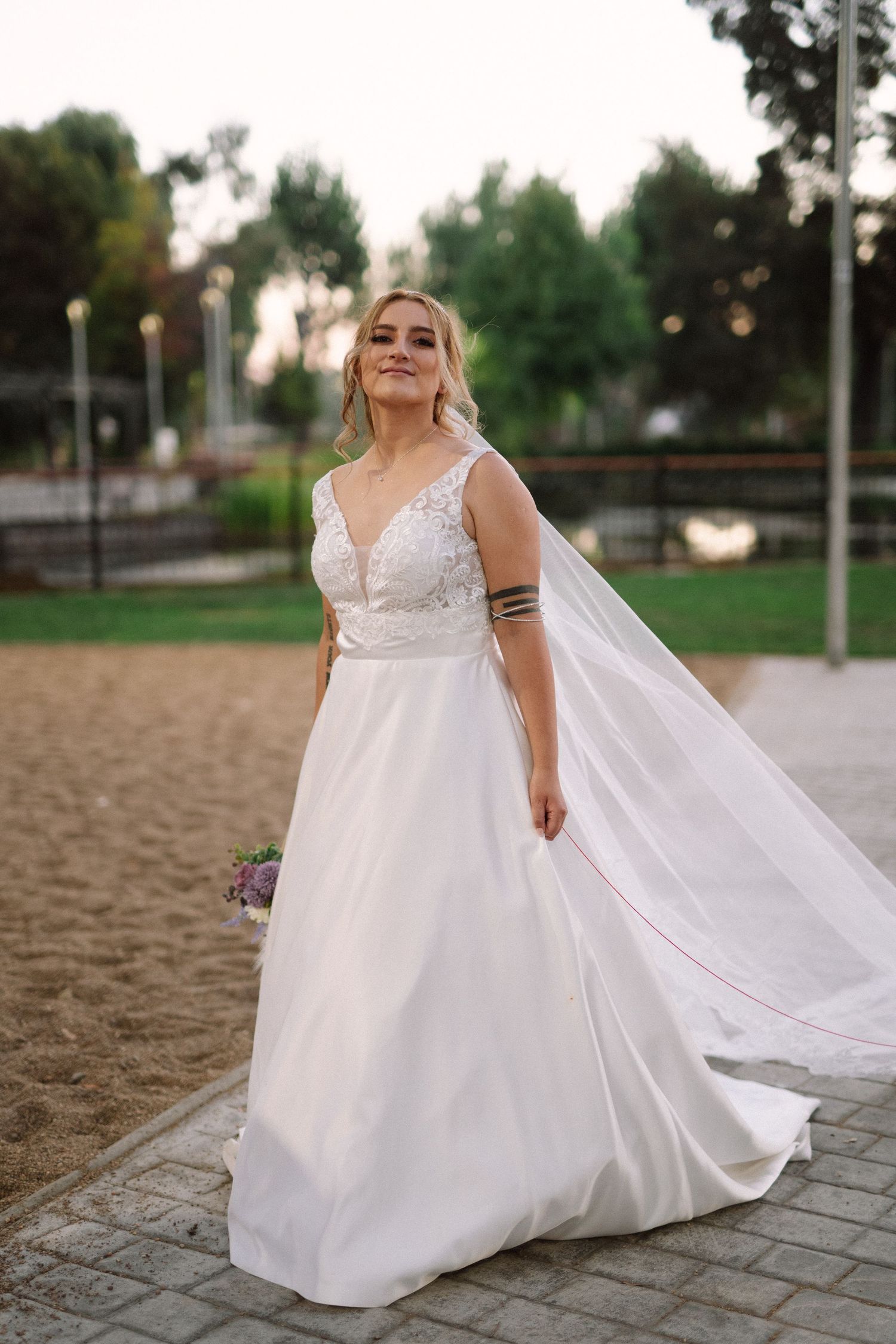 A bride in a white lace wedding gown with flowing veil poses outdoors at sunset on a paved path.