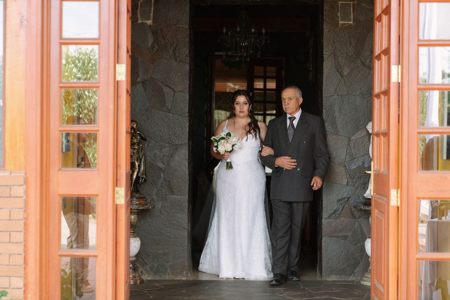 Bride in white gown holding bouquet walks with father through wooden doors at wedding ceremony entrance.