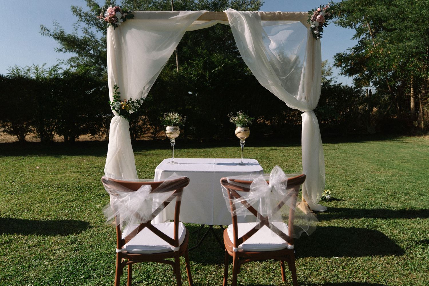 Elegant outdoor wedding ceremony setup with white draped canopy, floral arrangements, and two wooden chairs on green grass.