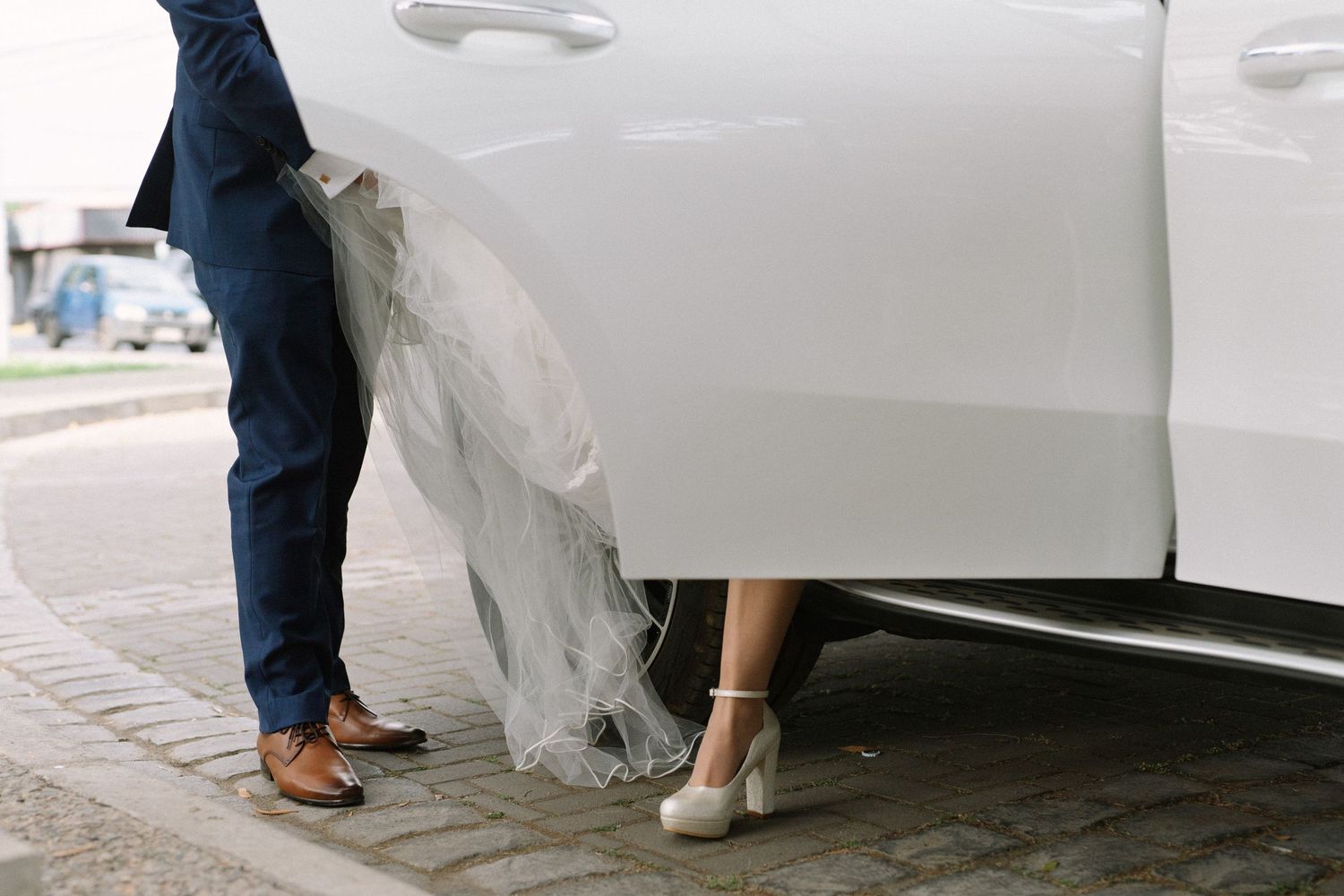 Detail shot of bride's dress and groom's shoes next to white car.