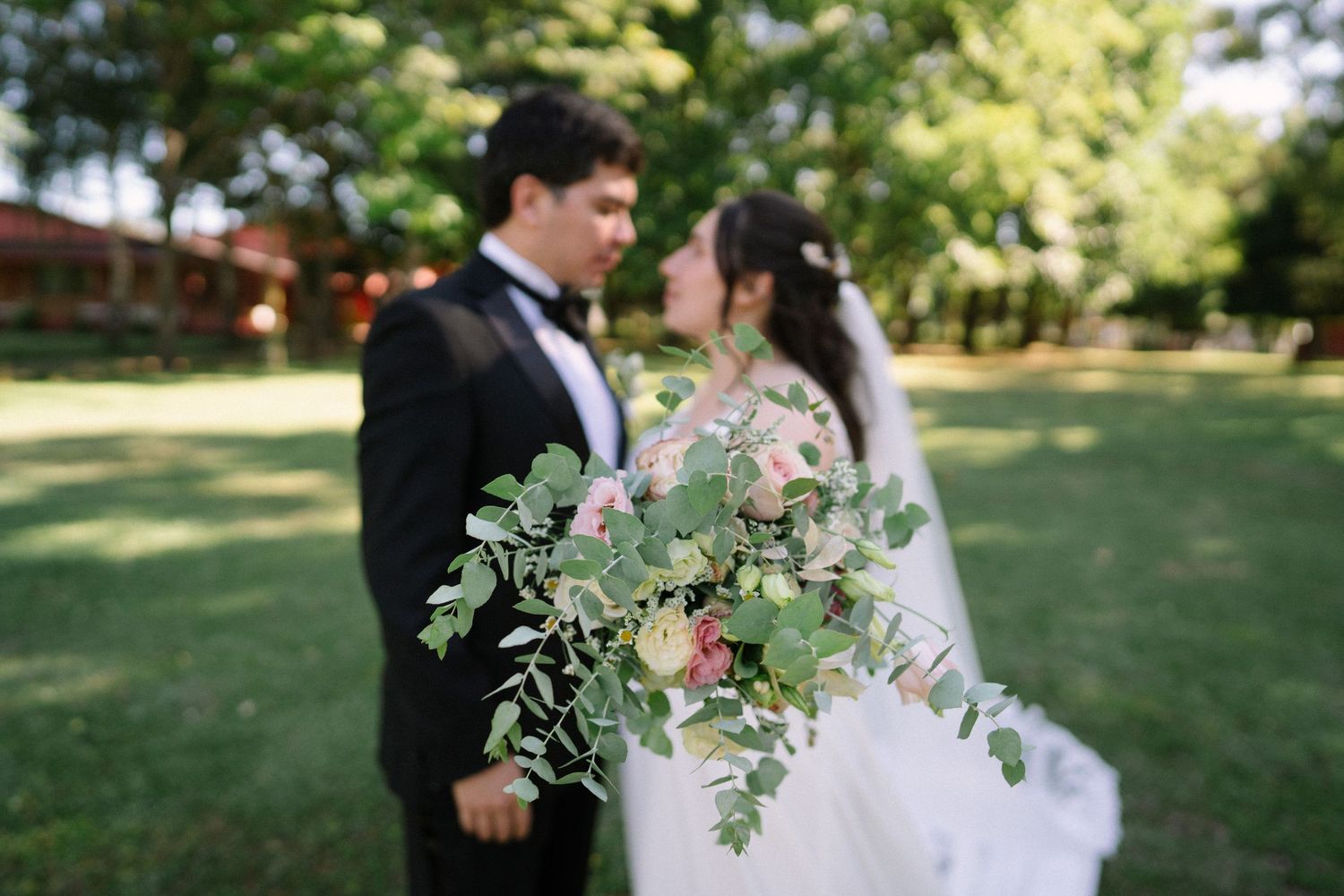 Newlyweds pose with a beautiful eucalyptus and rose bouquet in an outdoor garden setting.