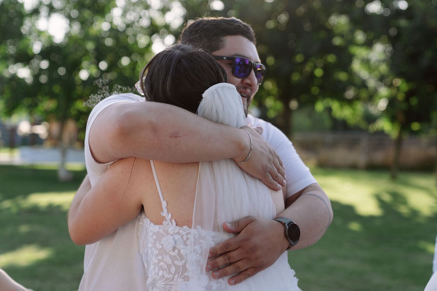 Emotional wedding guest hugs bride in white lace dress at outdoor garden ceremony.