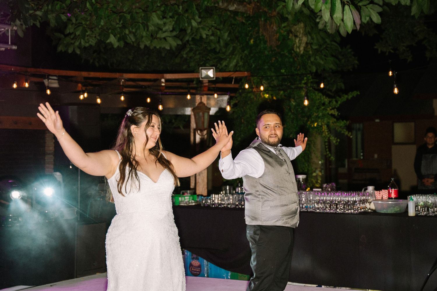 Bride and groom raise arms joyfully on dance floor at elegant outdoor evening wedding reception.
