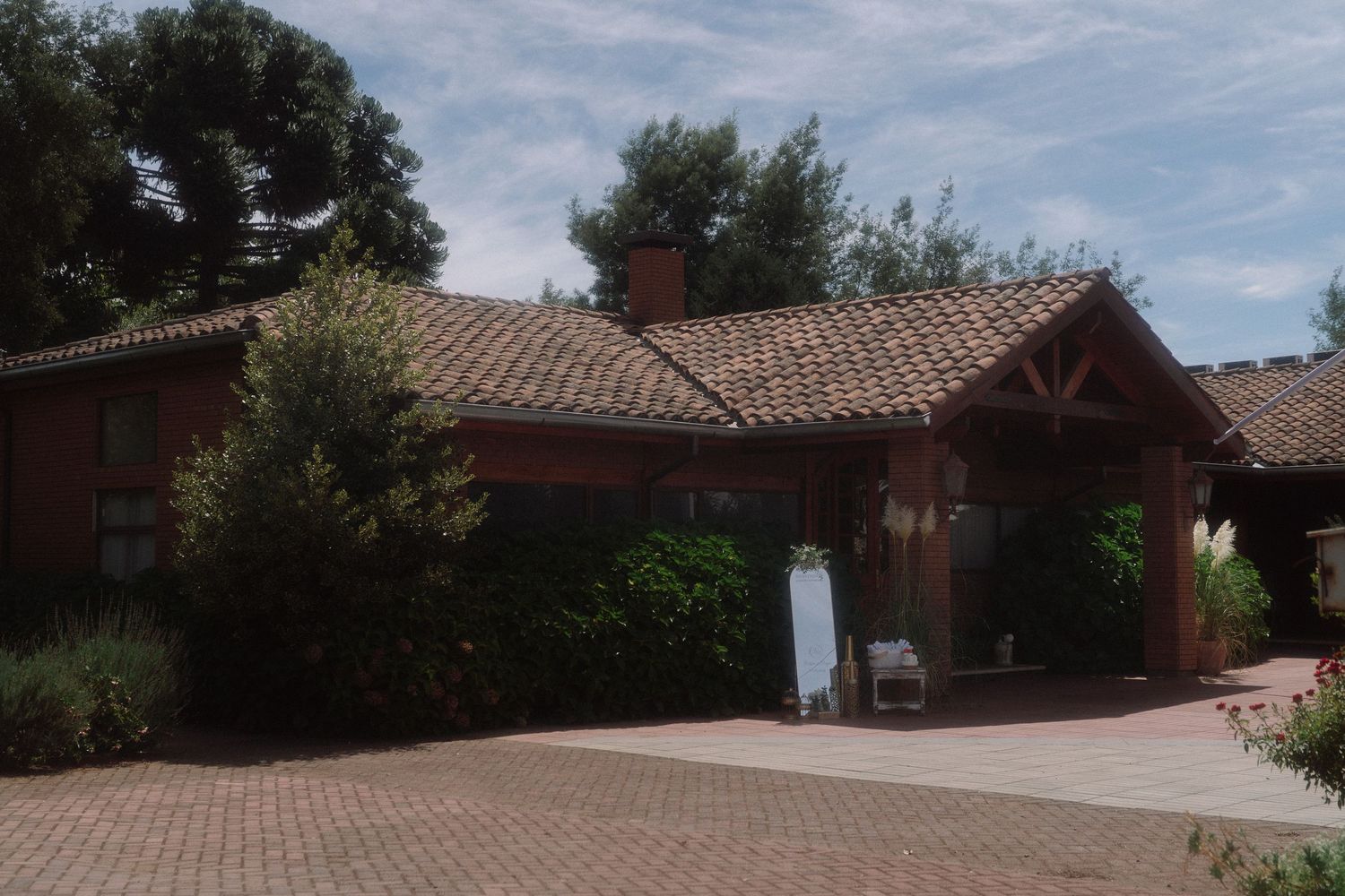 Red ranch-style home with terracotta tile roof, brick driveway, and lush green trees in a warm sunny setting.