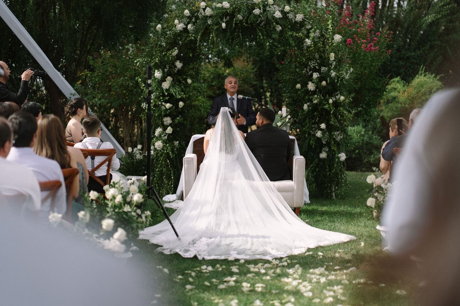 Officiant addresses bride and groom during an outdoor garden wedding ceremony with white floral arch.