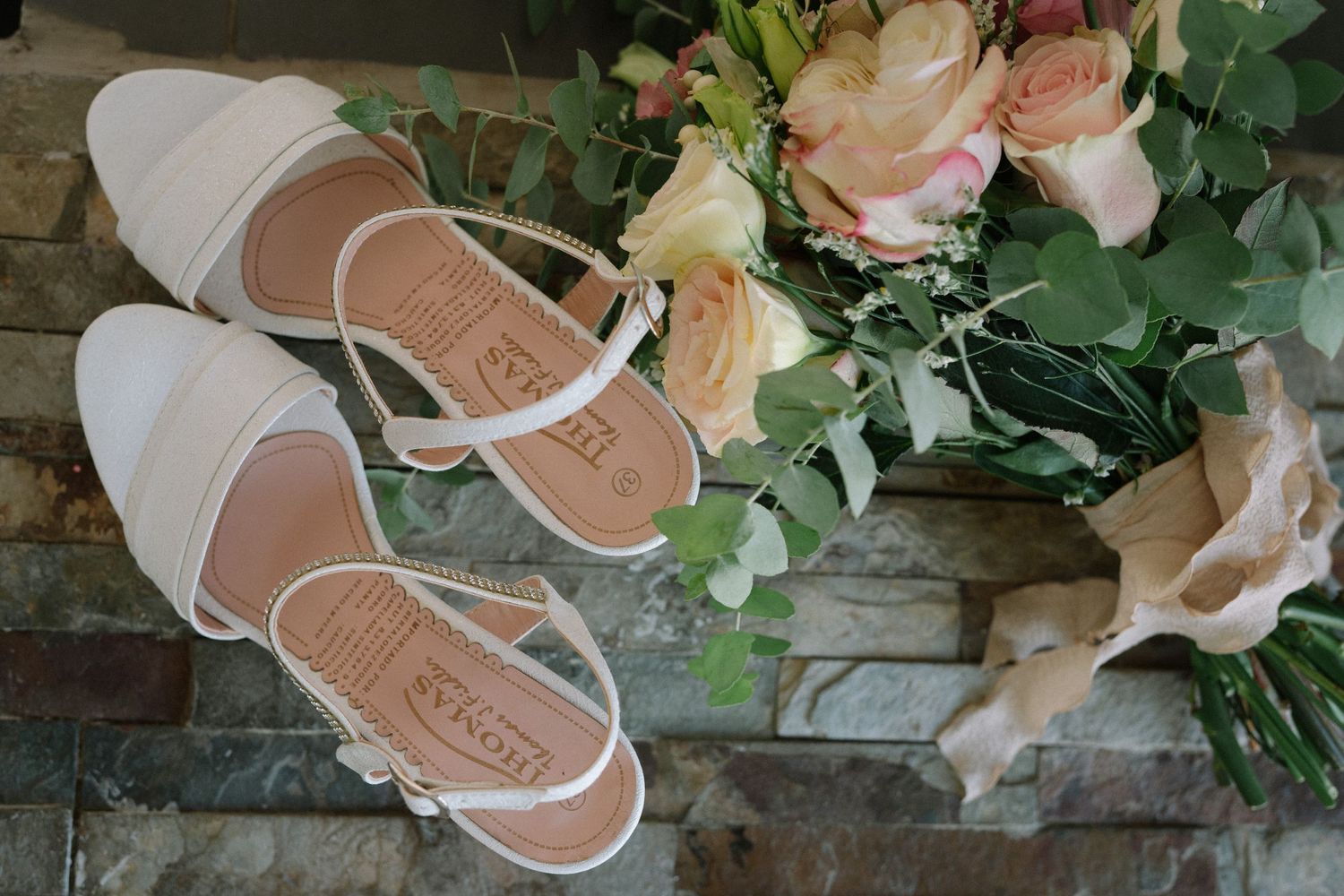 White bridal sandals arranged with pink roses and greenery on a stone surface.
