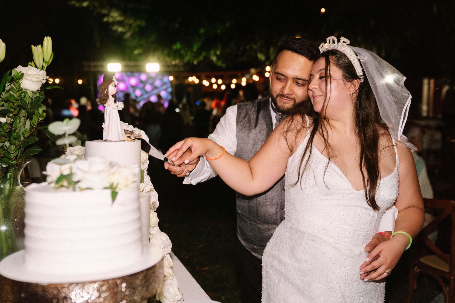 Newlywed couple cutting white tiered wedding cake together at outdoor evening reception celebration.