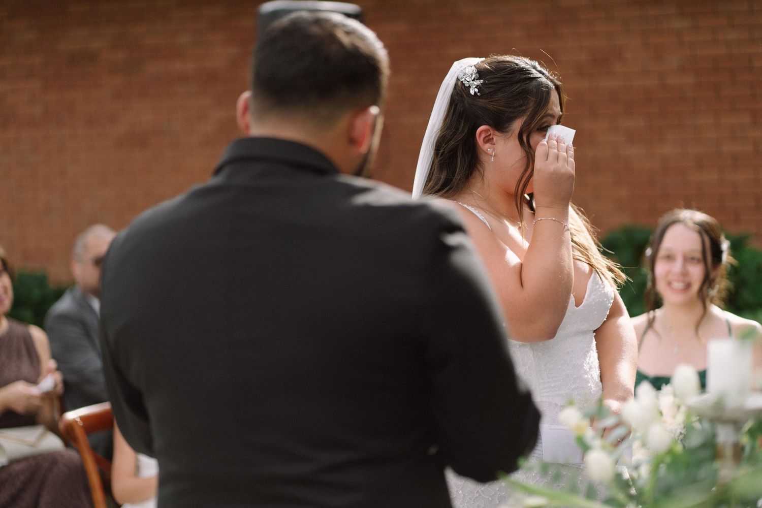 Emotional bride wipes tears during outdoor wedding ceremony while groom stands before her.
