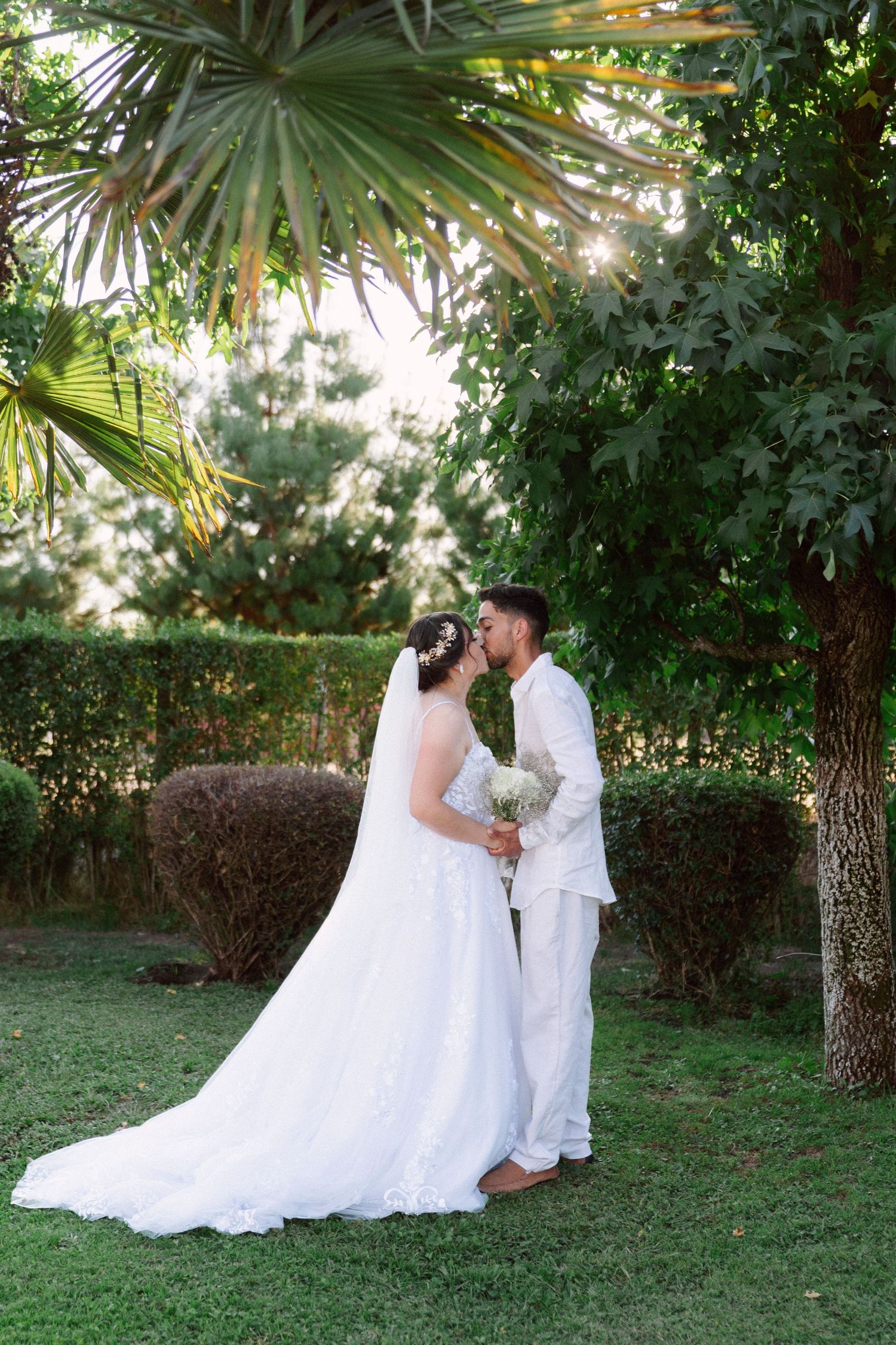 Bride and groom share a romantic kiss under lush palm trees in a sunlit garden after their wedding.