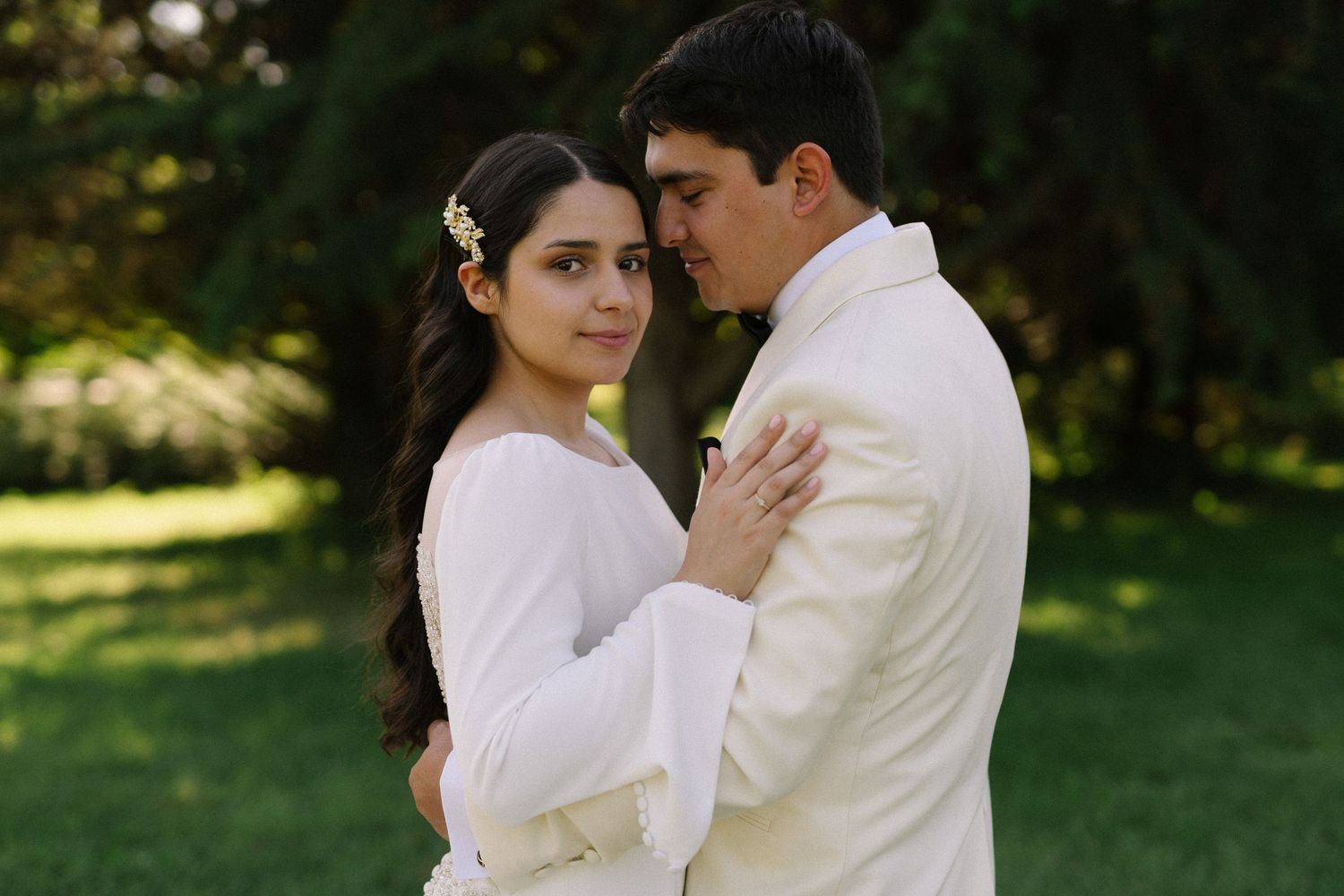 A romantic outdoor wedding portrait of a couple in white outfits embracing in a sunlit garden setting.
