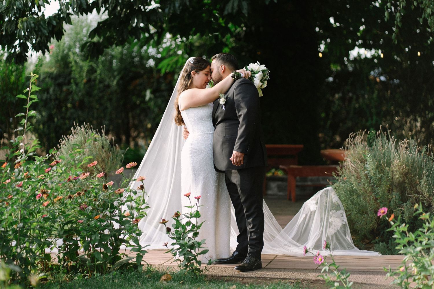 A bride and groom share their first kiss surrounded by lush garden greenery on their wedding day.