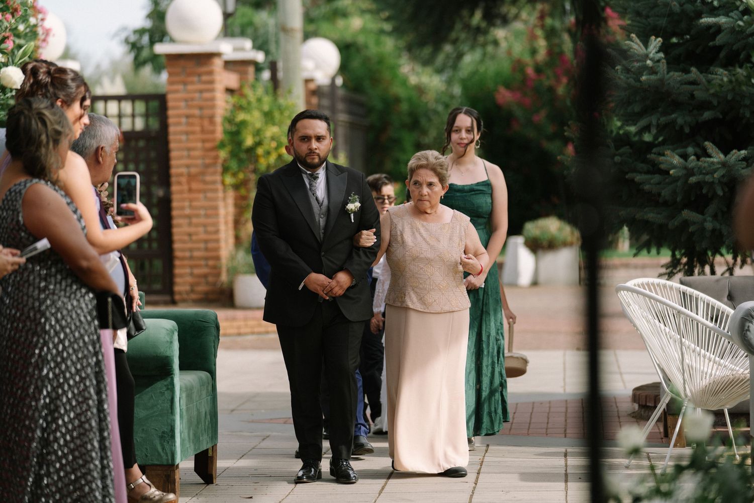 Groom in black suit escorts elderly woman down outdoor wedding aisle flanked by guests.