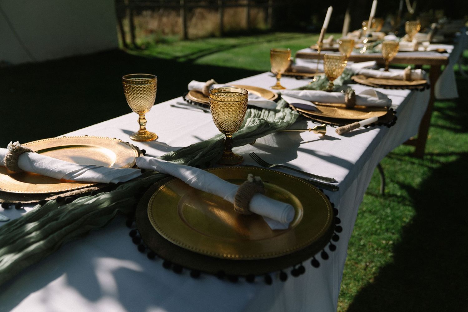 Elegant outdoor table setting with gold charger plates, amber glassware, and white linen napkins on a white tablecloth.