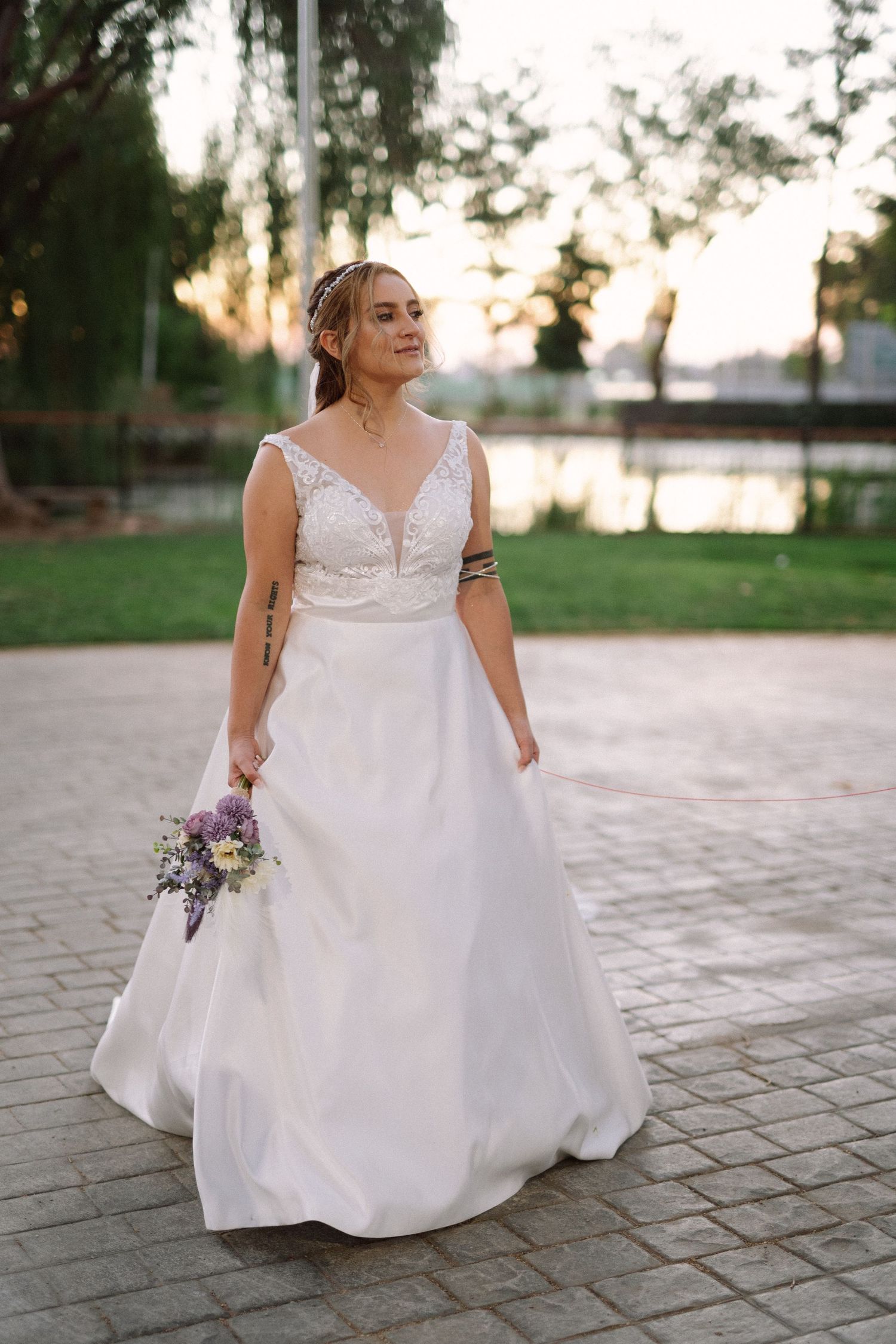 A bride in a V-neck wedding gown holds a purple bouquet against a sunset backdrop.