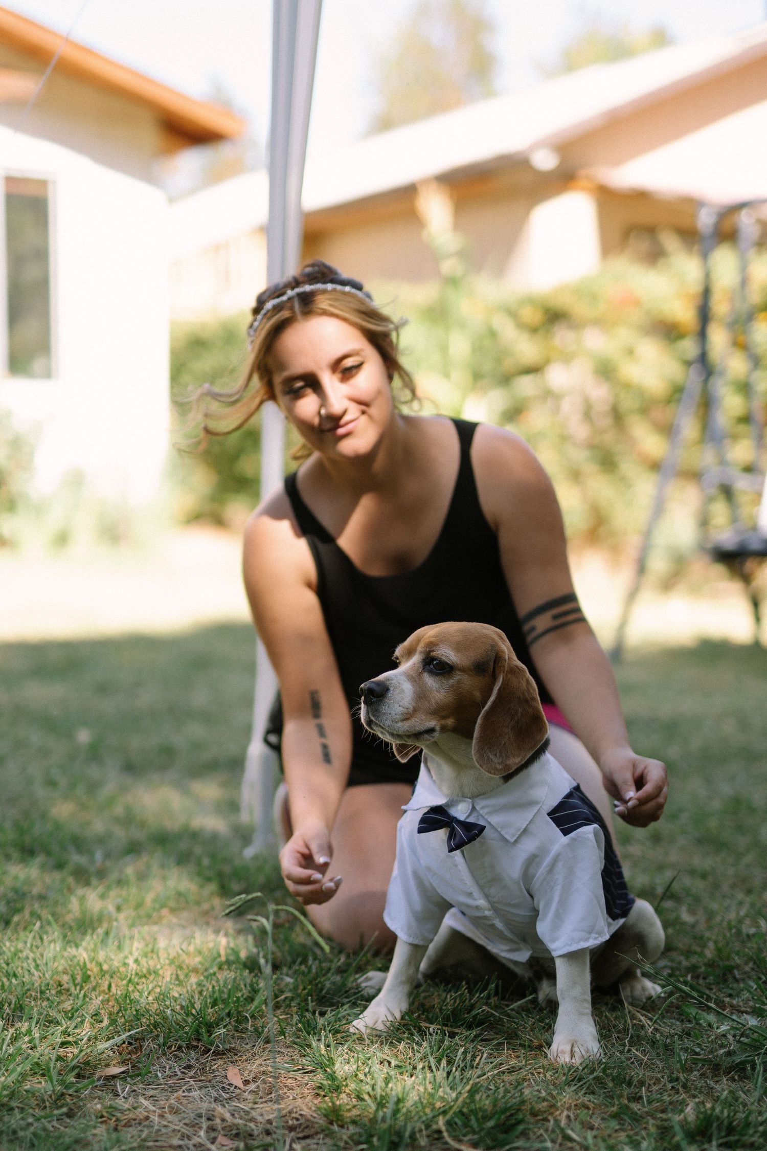 A beagle wearing a formal white shirt sits with someone on grass in a backyard setting.