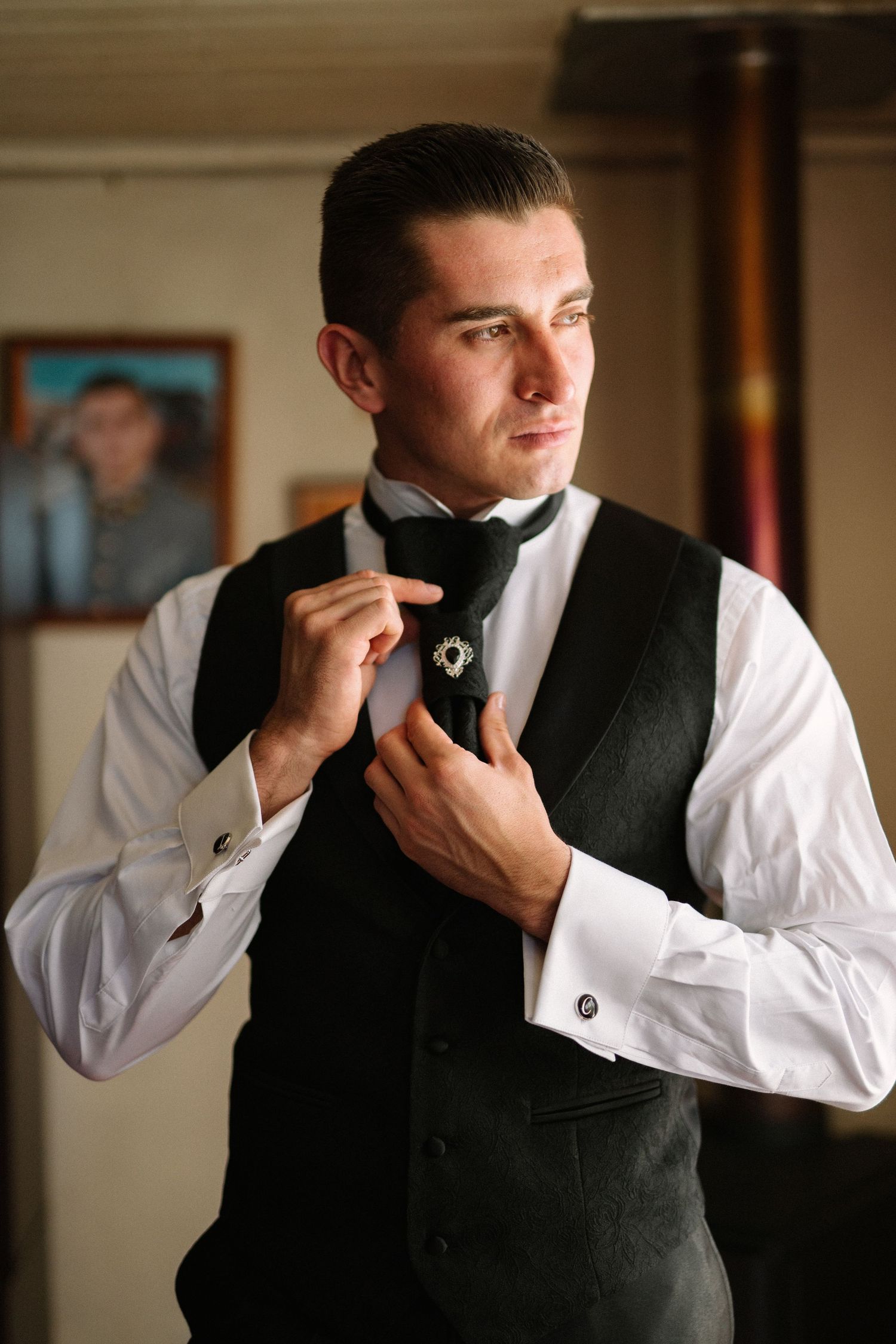 A man in formal attire adjusts his bow tie while wearing a dark vest and white dress shirt.