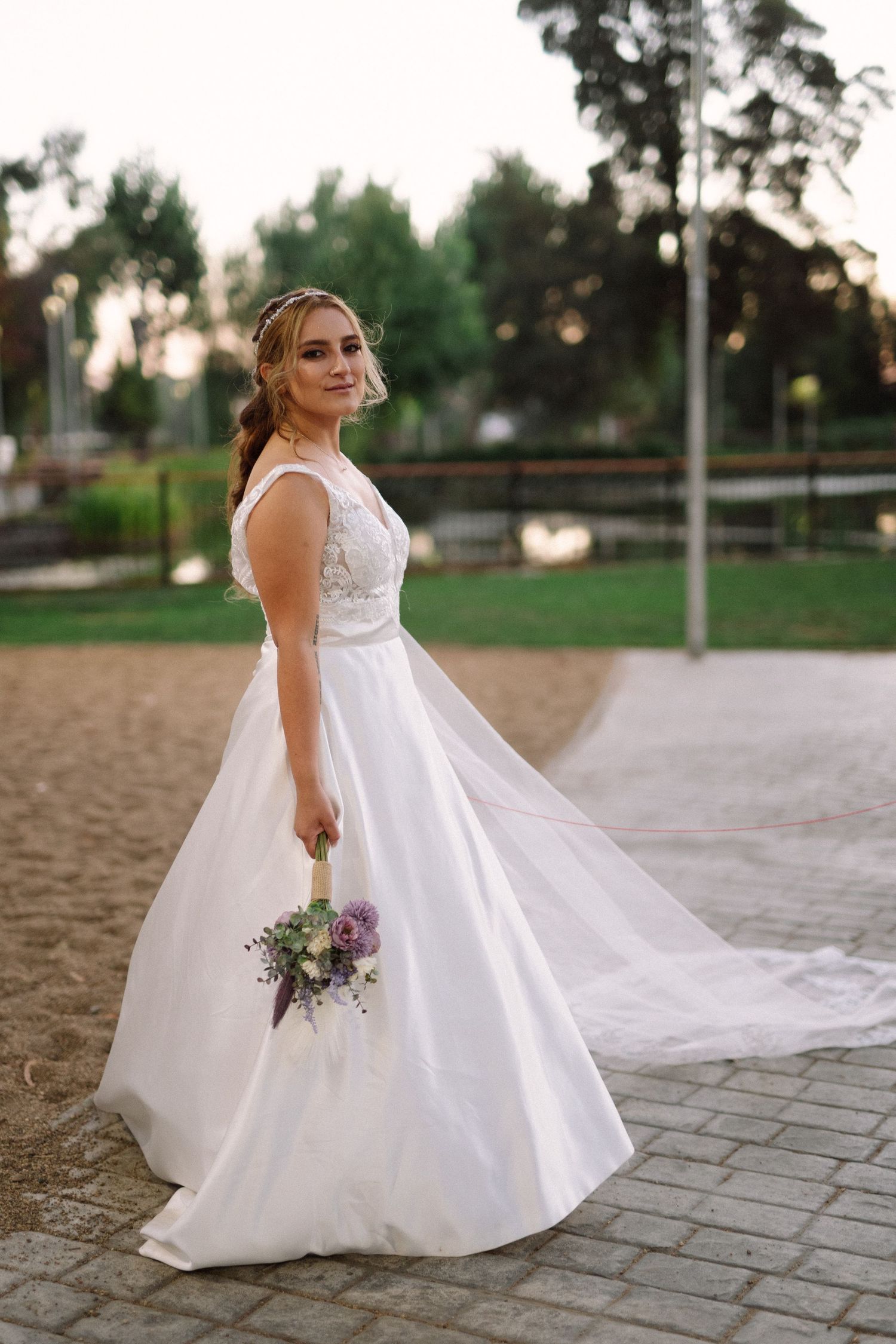 A bride in a flowing white dress poses with a purple bouquet on a paved path.
