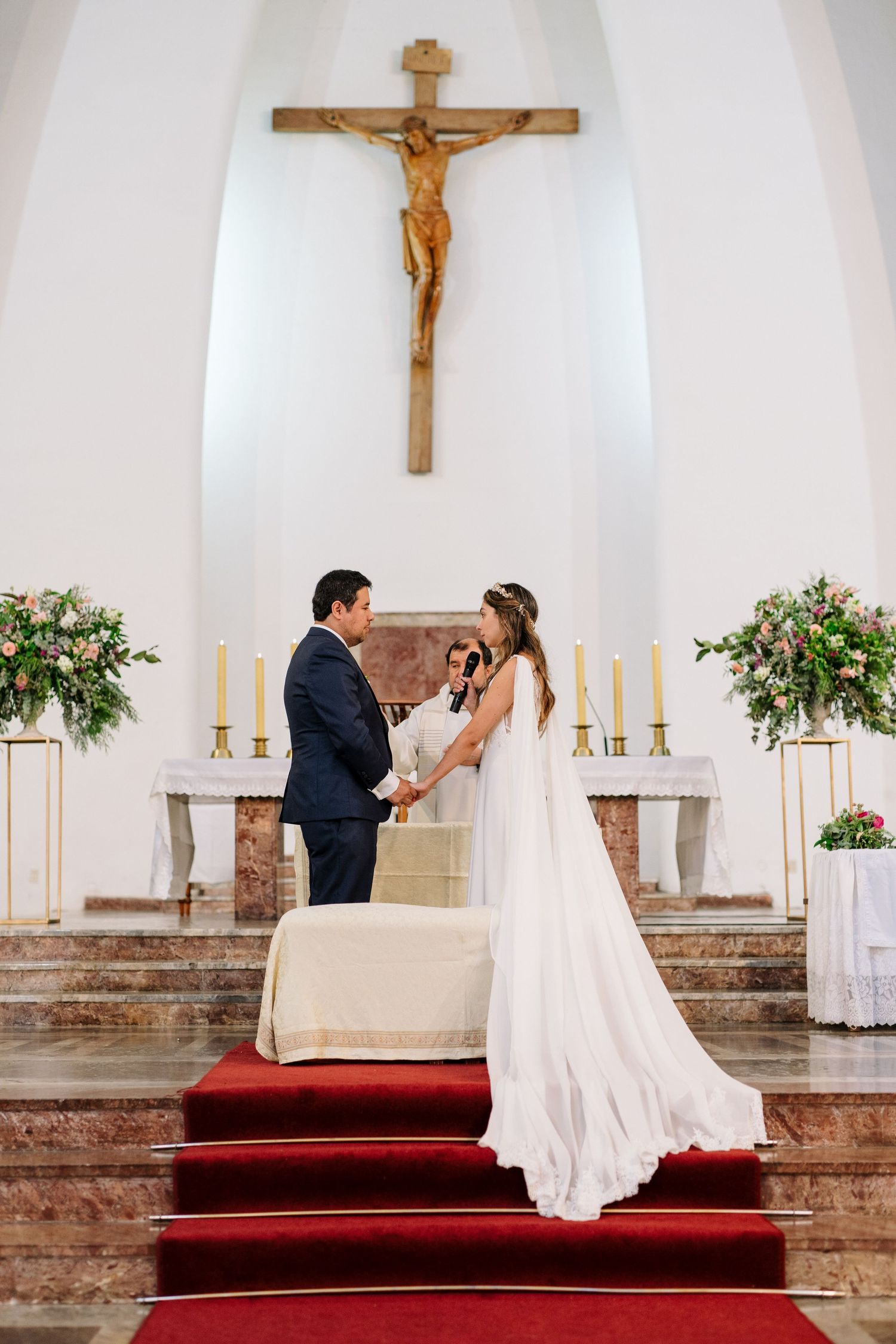 A couple stands under a crucifix on red-carpeted church steps during their wedding ceremony.
