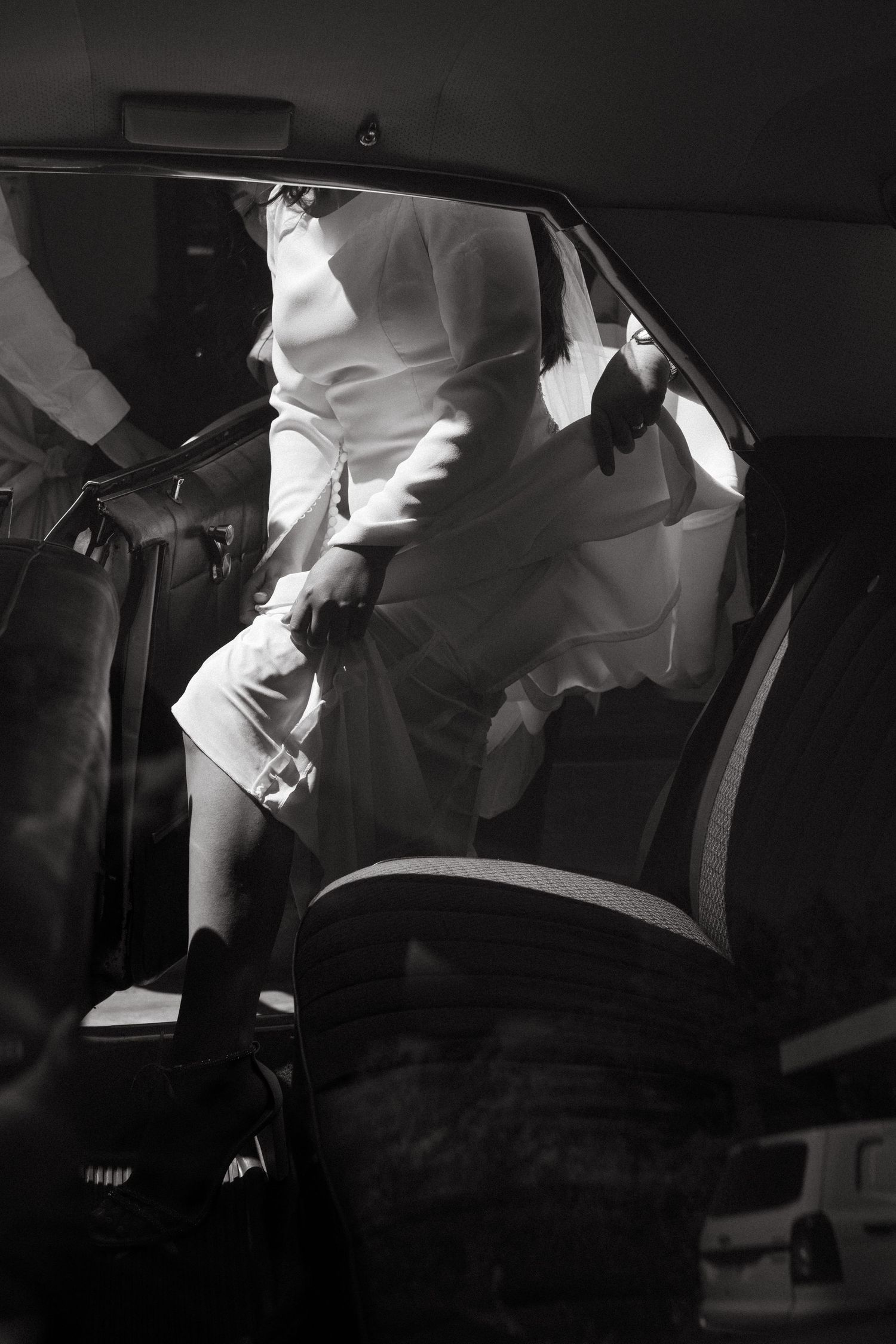 Black and white photo of a bride in white dress getting into a car.