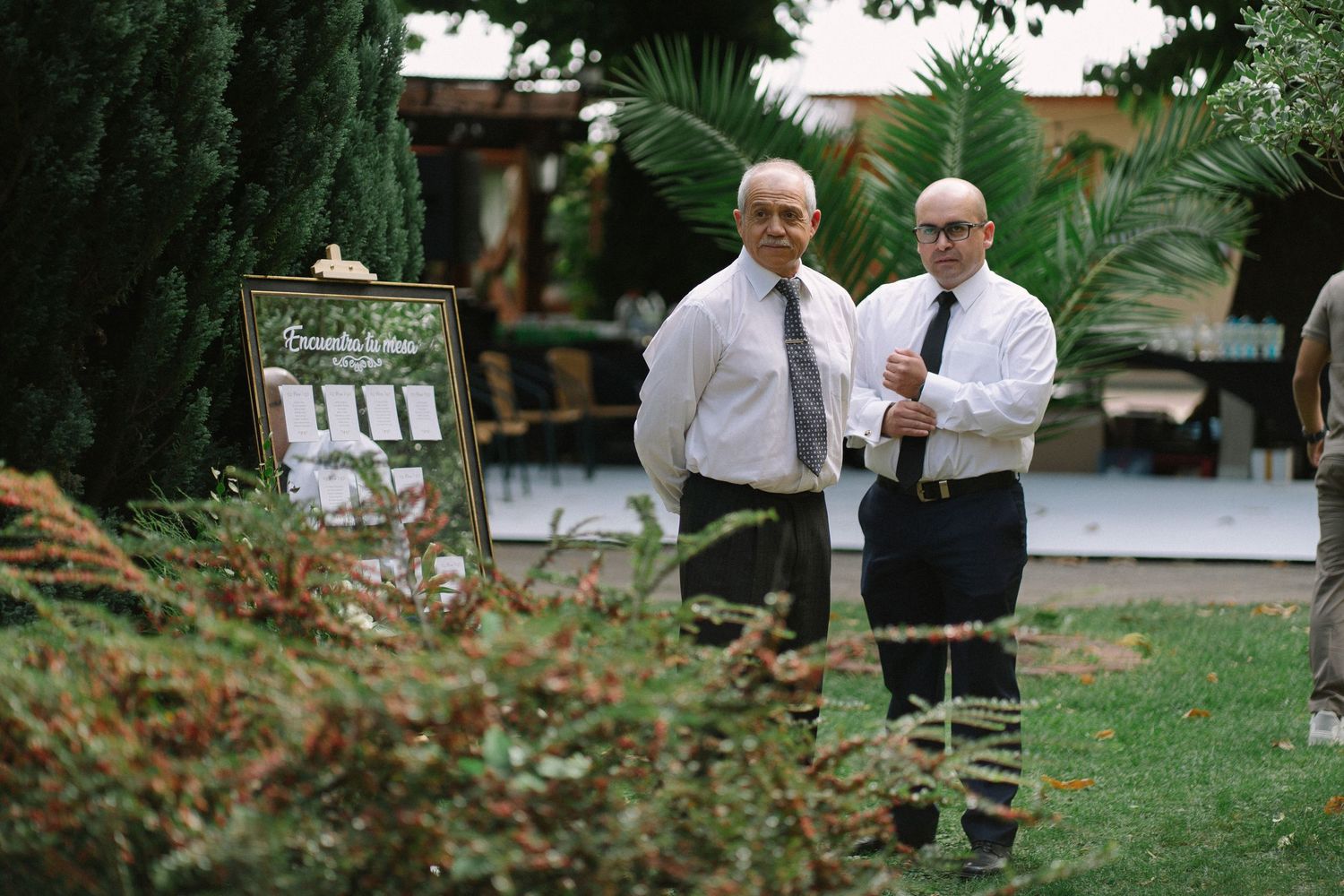 Two men in white shirts and ties standing outdoors near a seating chart sign at a garden wedding venue.