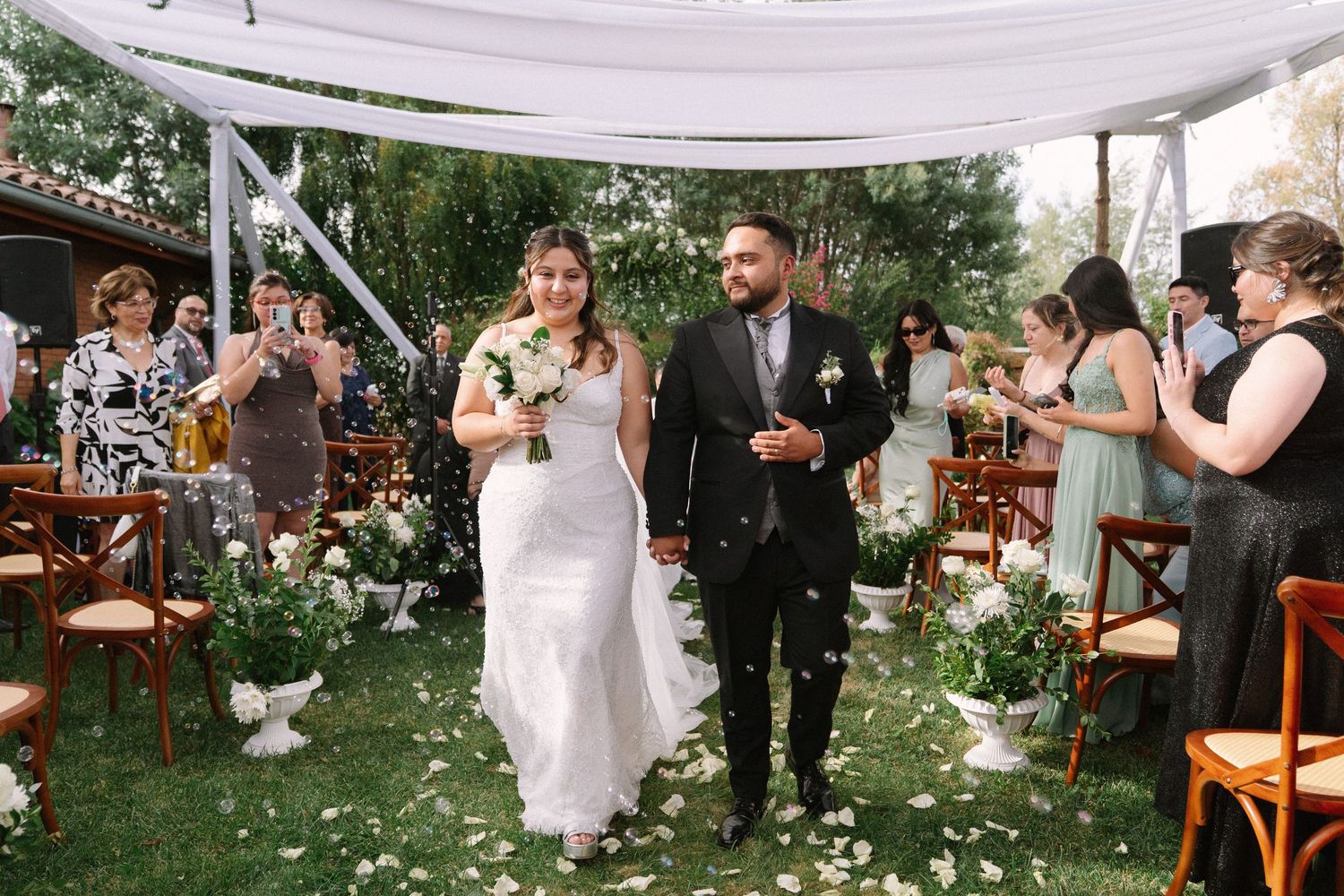 Newlyweds walk down flower petal aisle as guests celebrate with confetti after ceremony.