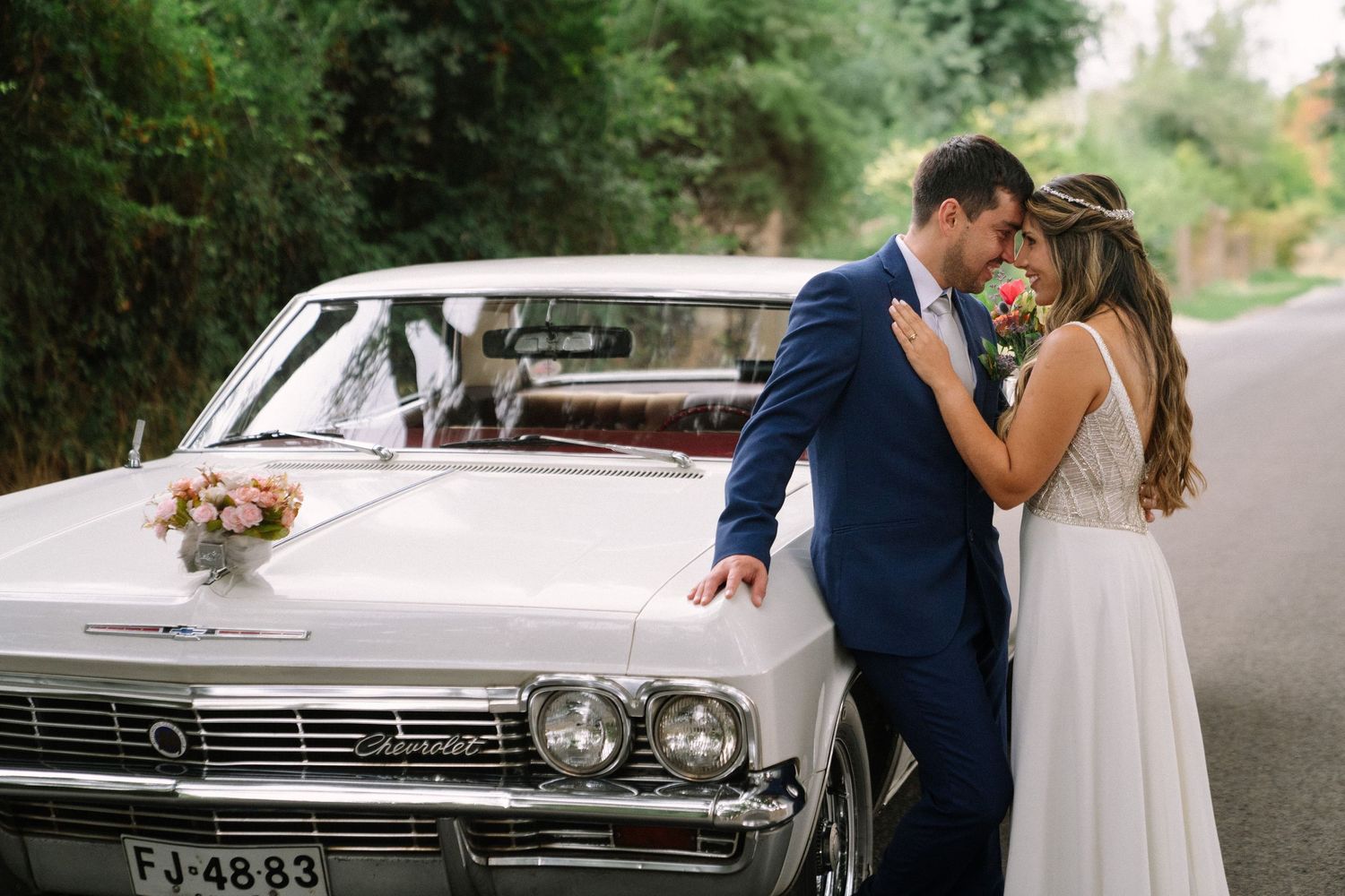 A romantic couple poses with a vintage white Chevrolet car decorated with flowers on a tree-lined road.