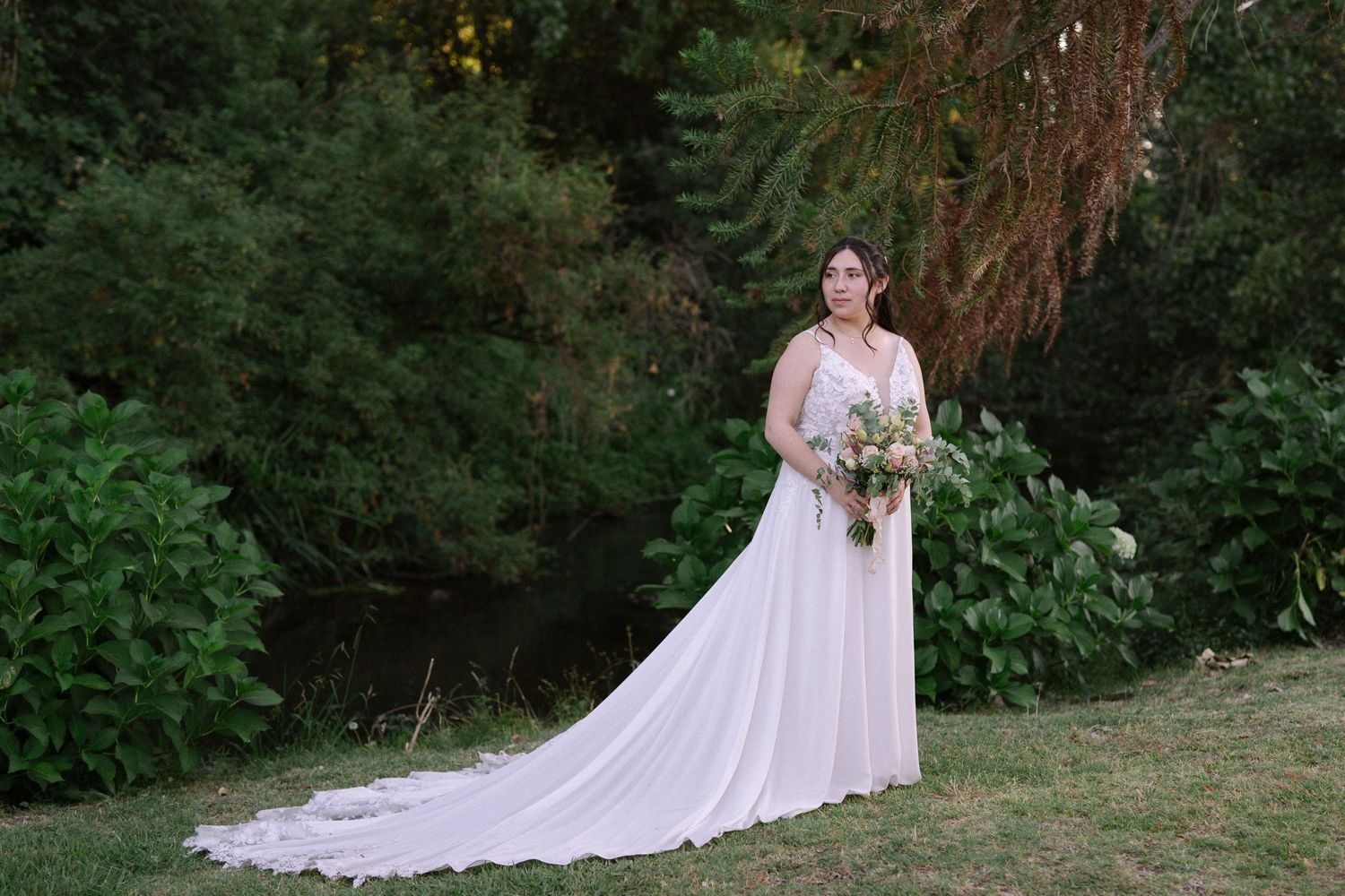 A bride in a flowing white dress poses in a garden setting with a rustic backdrop.