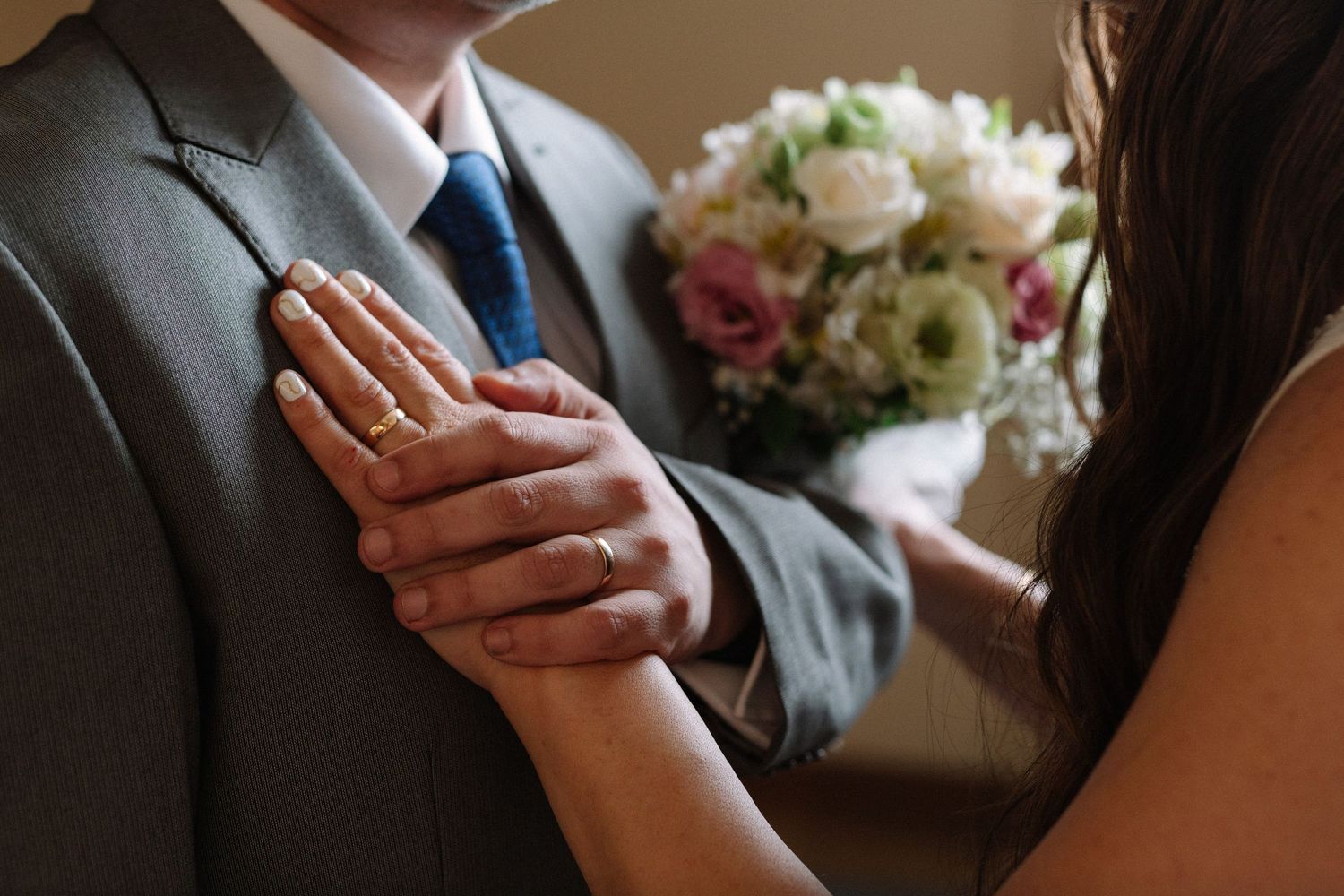 Intimate moment showing clasped hands and wedding rings with bouquet in background.