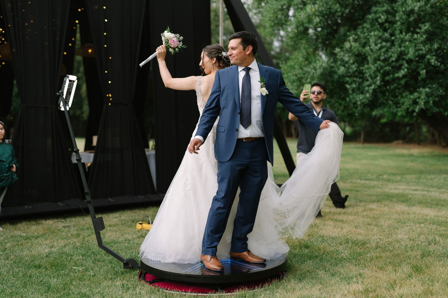 A couple in wedding attire dances playfully on a large tire swing in an outdoor garden setting.