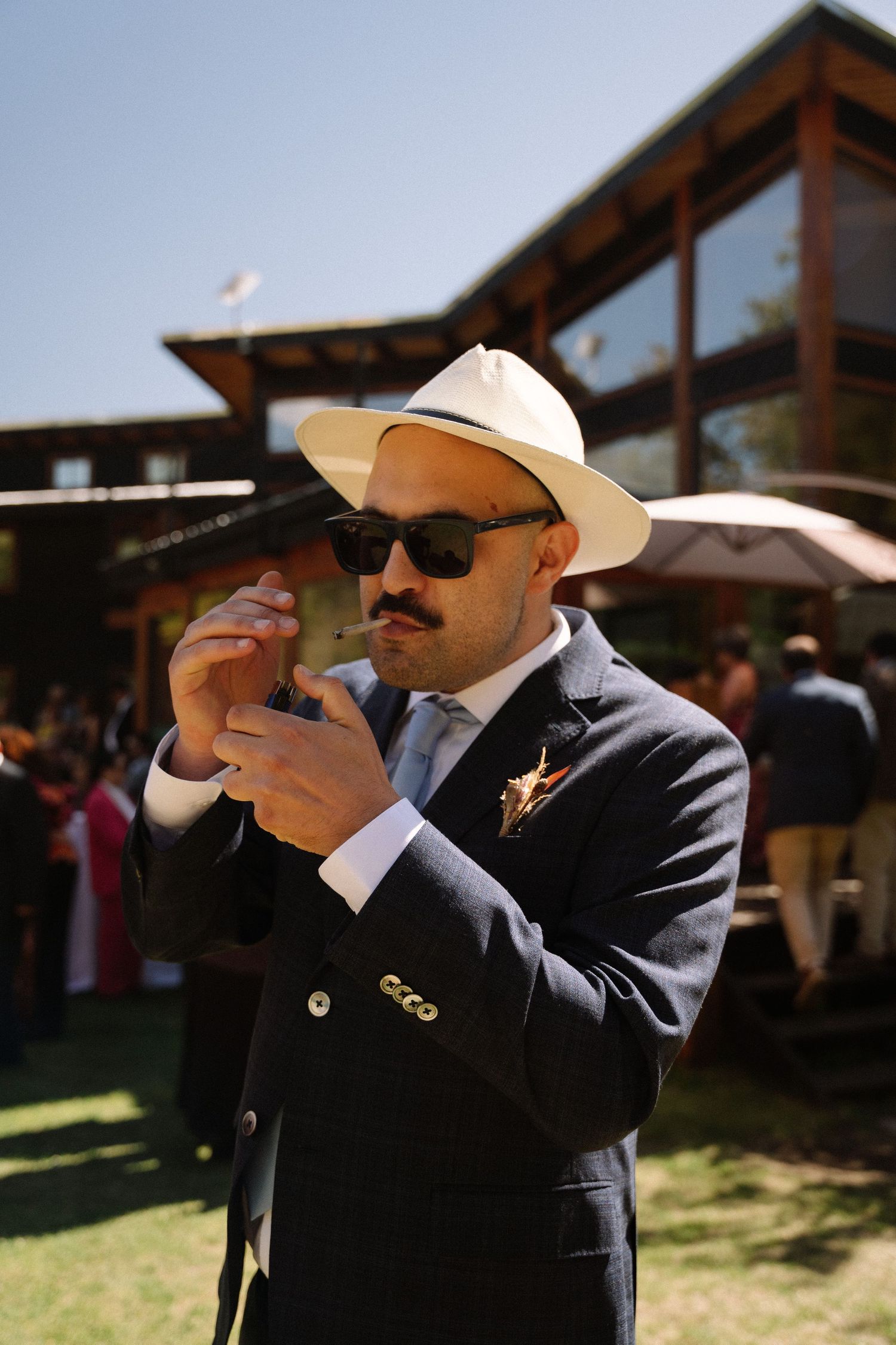 A well-dressed guest in a suit and cowboy hat lights a cigar at an outdoor wedding.