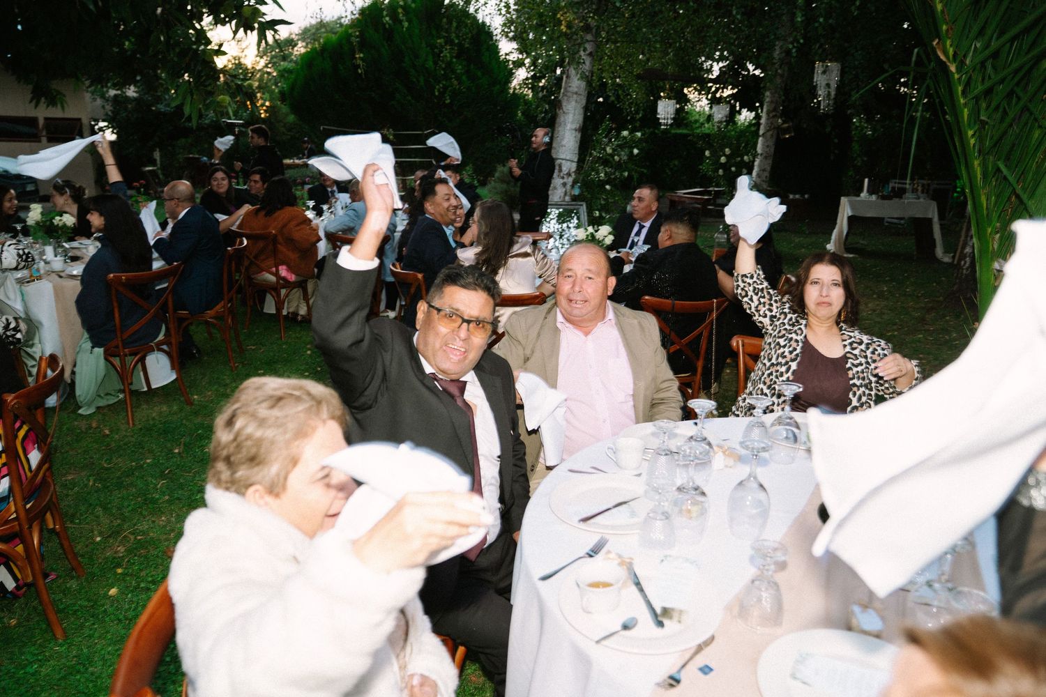 Wedding reception guests waving white napkins in celebration at elegantly decorated outdoor tables.