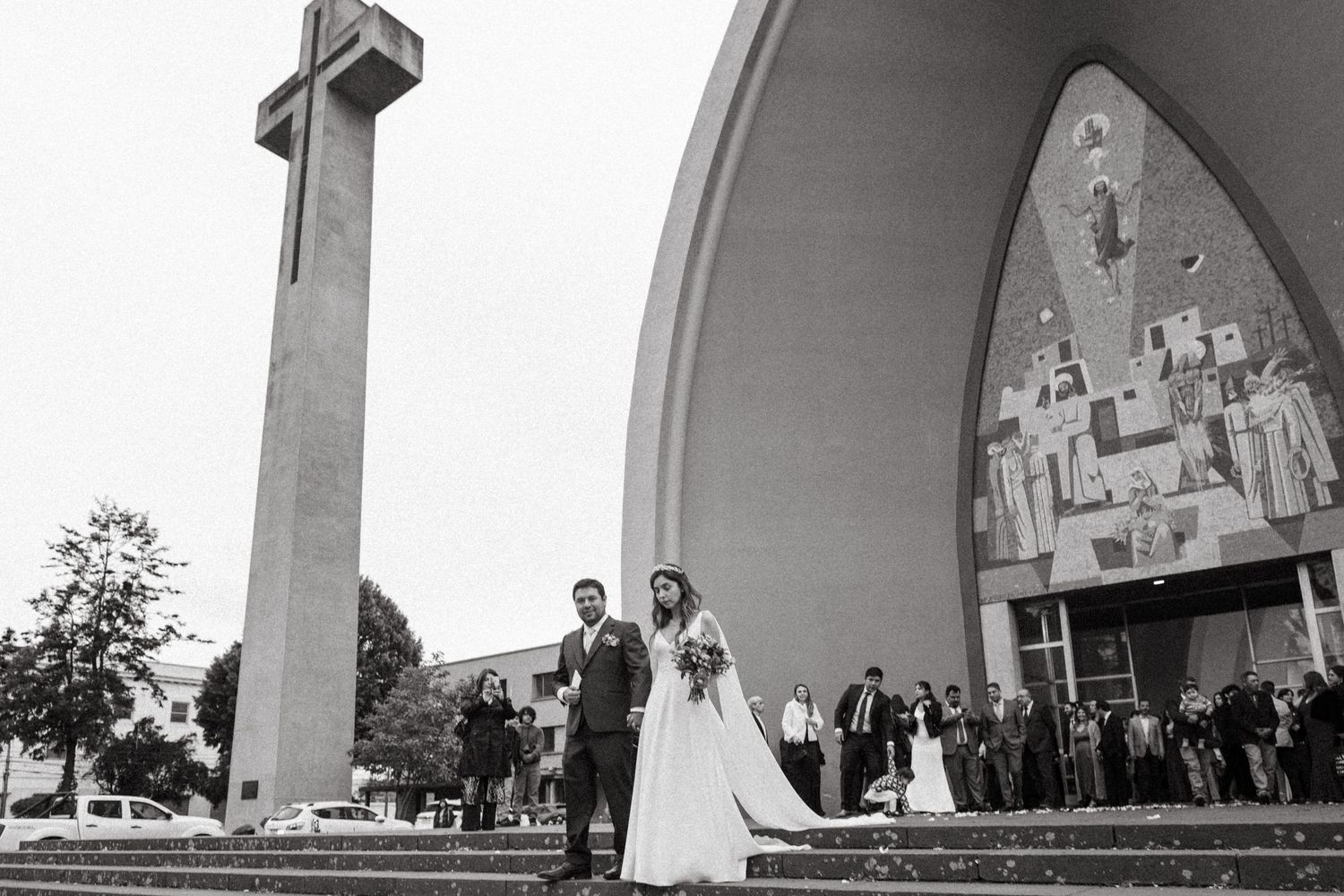 Black and white photo of wedding couple outside modern church with tall cross monument.