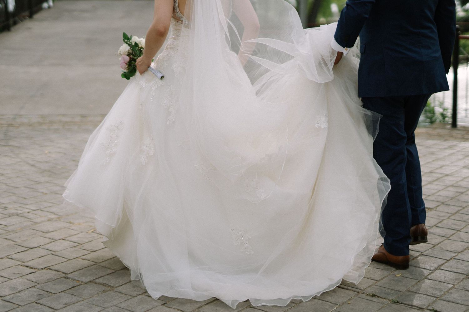 A bride's flowing white wedding dress billows as she walks along a stone path.