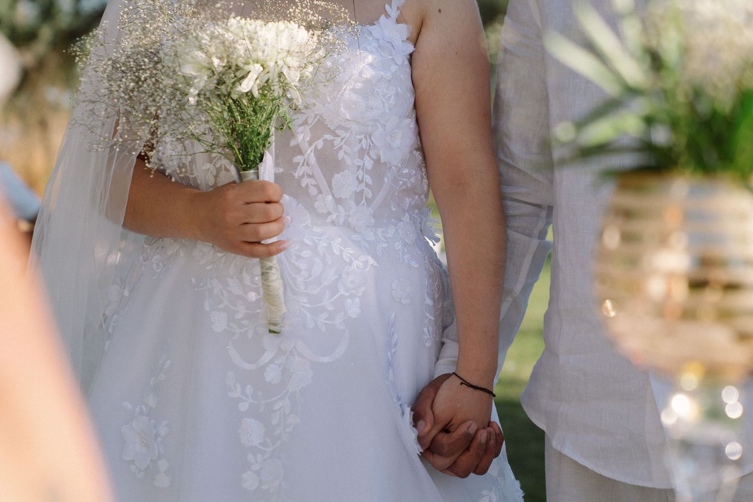 Bride in lace wedding dress holding white bouquet while holding hands with groom at outdoor ceremony.