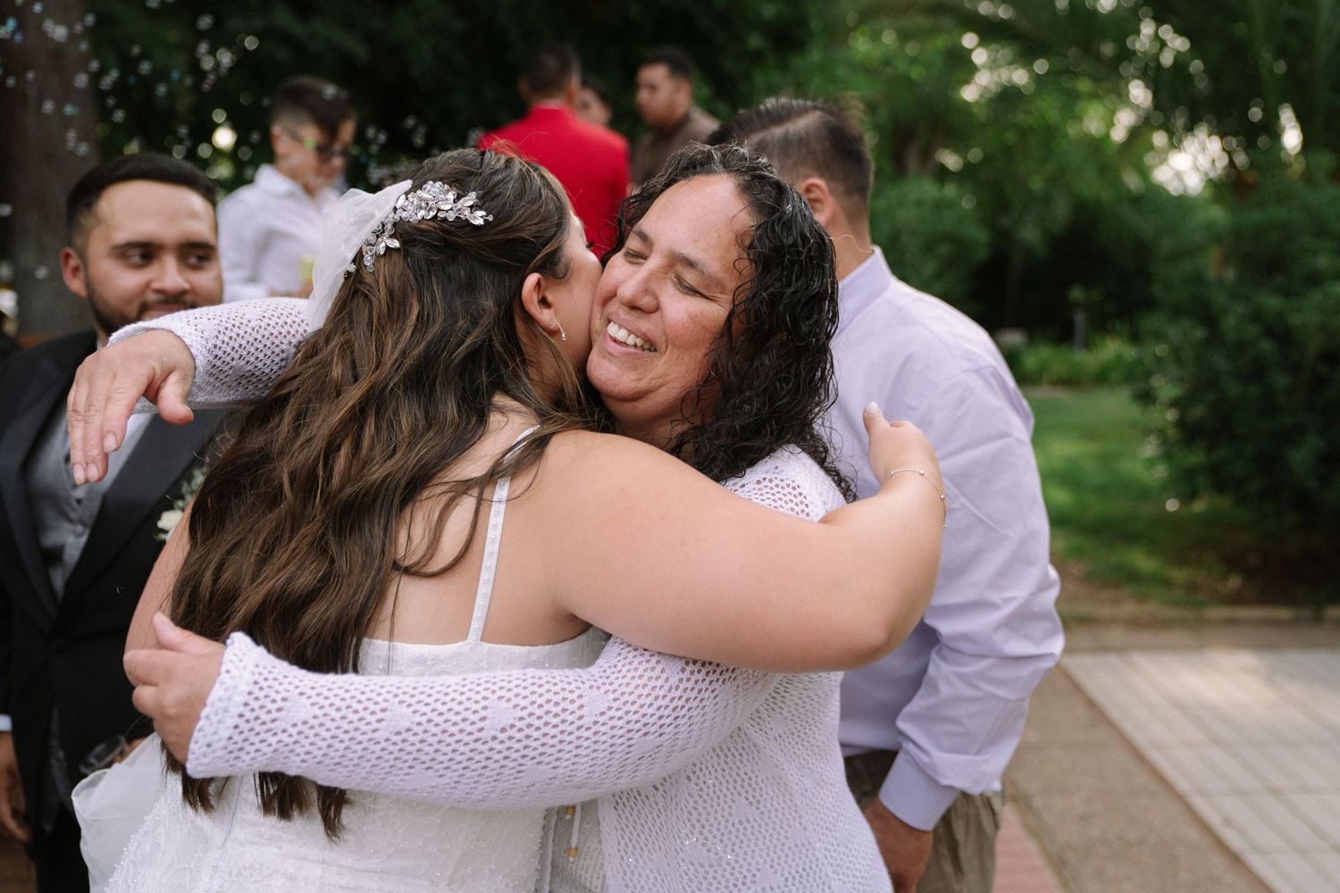 Bride in white wedding dress shares warm embrace with smiling female guest at outdoor wedding reception.