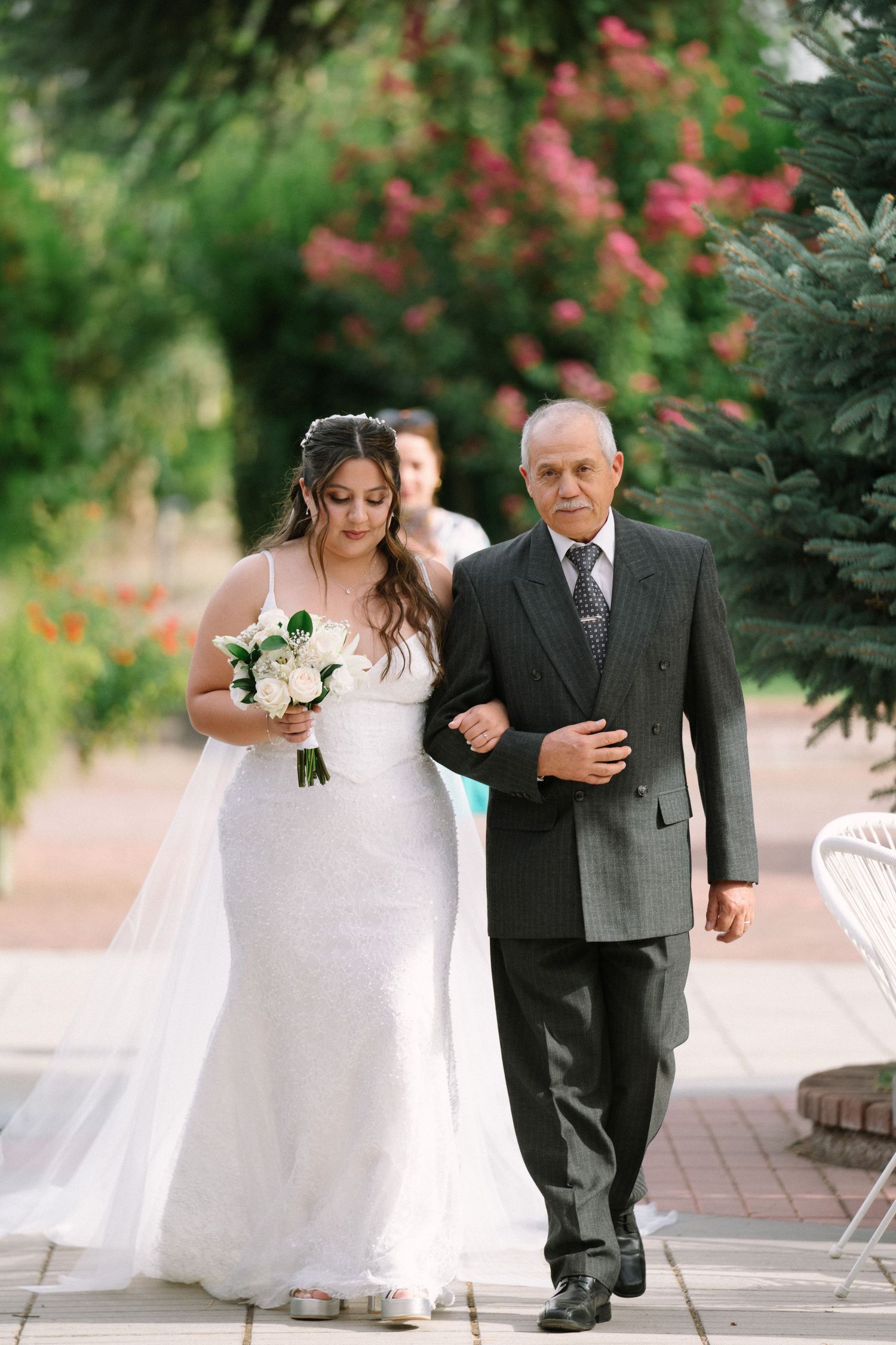Bride in white gown holding bouquet walks down aisle with older man in gray suit at outdoor wedding.